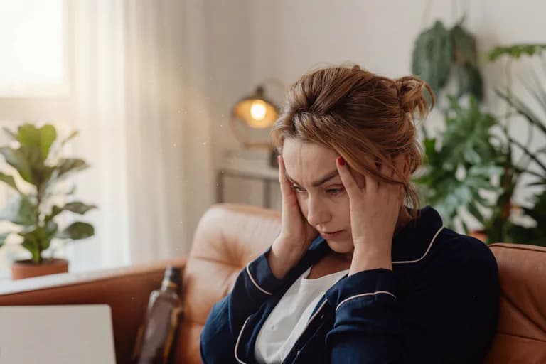 Kvinna med händerna mot tinningarna, ser ut att ha ont i huvudet eller vara stressad, inomhus i ett vardagsrum.