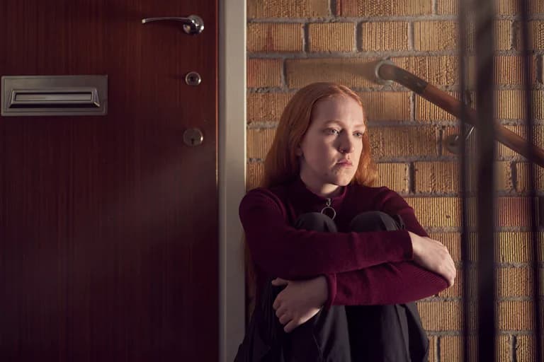 Kvinna med rött hår sitter hopkrupen på trappsteg framför en tegelvägg och tittar bort.