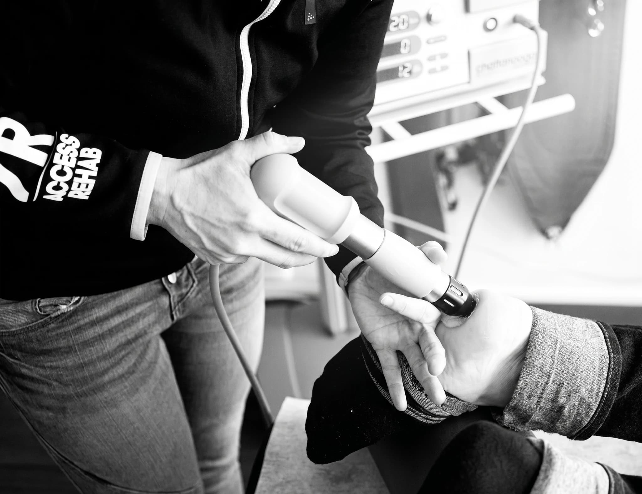 En terapeut använder stötvågsbehandling på en patients hand, med enheter för rehabilitering i bakgrunden.
