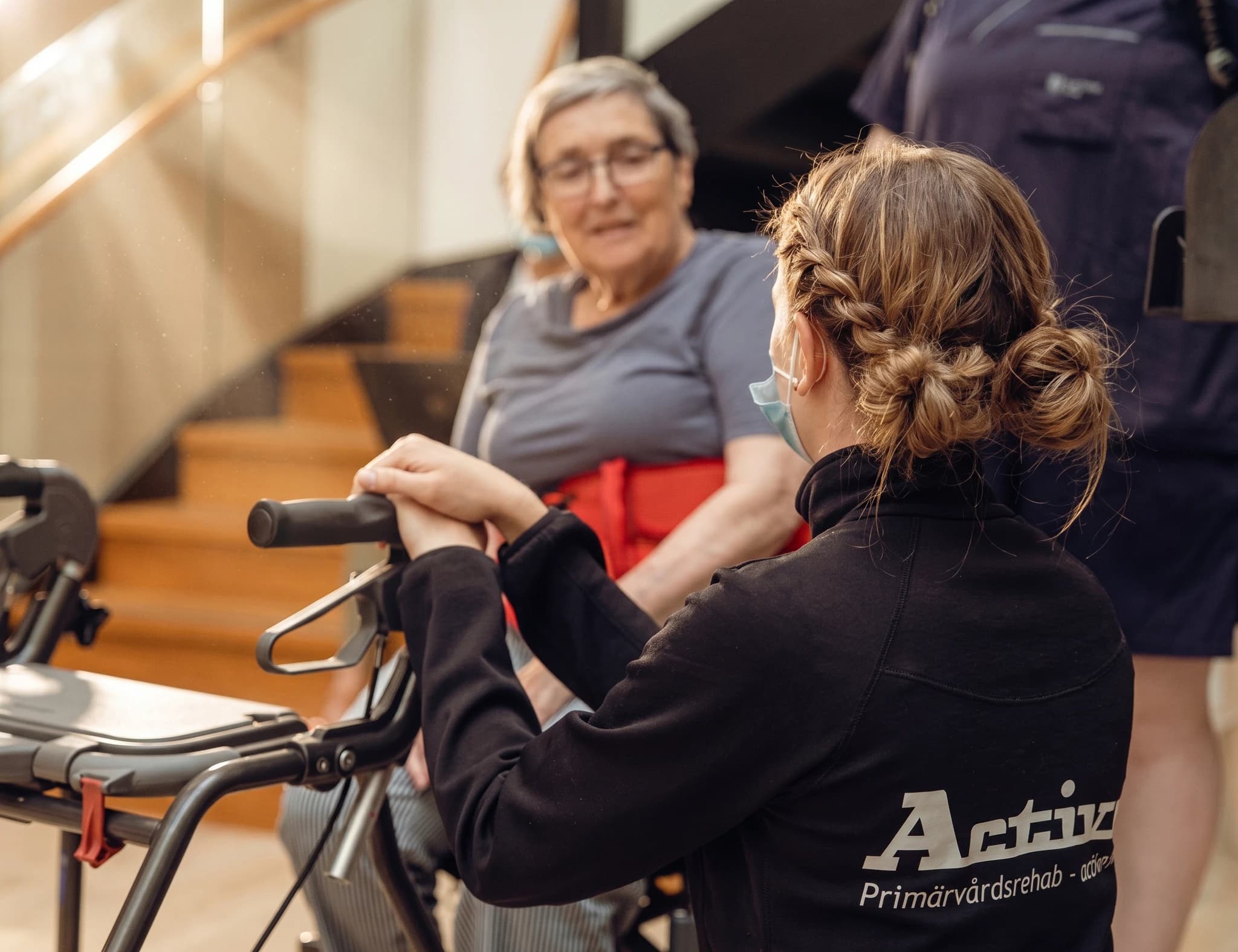 En terapeut med flätat hår hjälper en äldre kvinna med glasögon att använda en rollator.