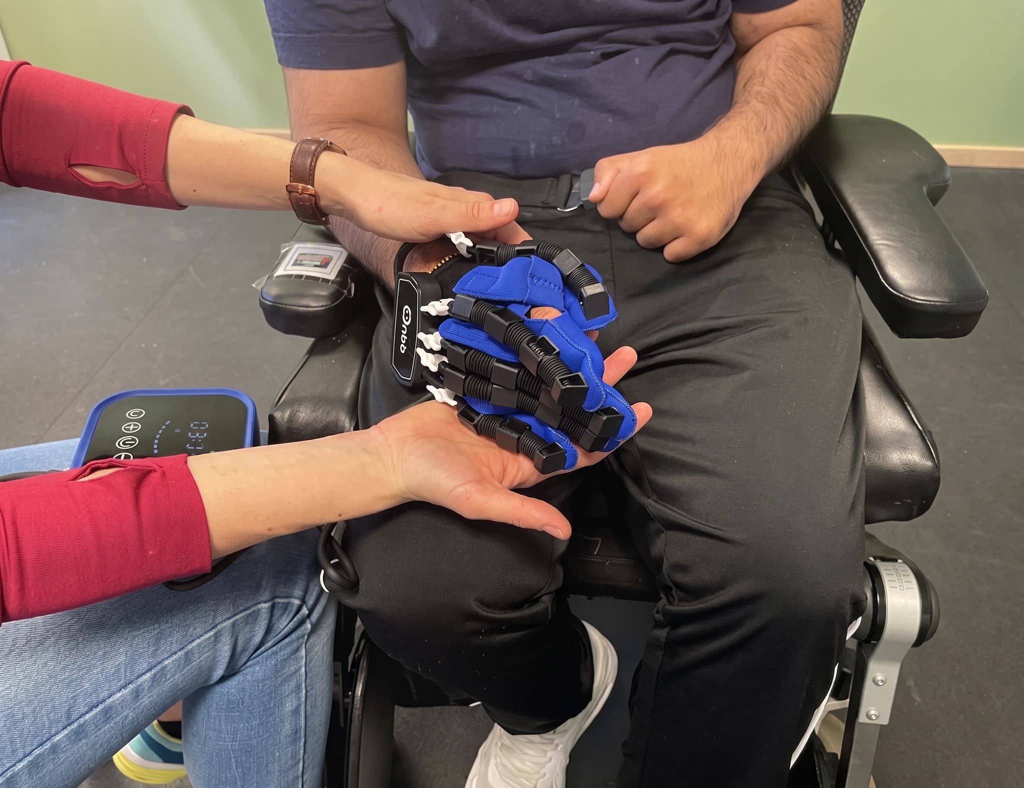 En person sitter i rullstol och får hjälp att trä på sig en robotiserad handske som en del av rehabiliteringen.