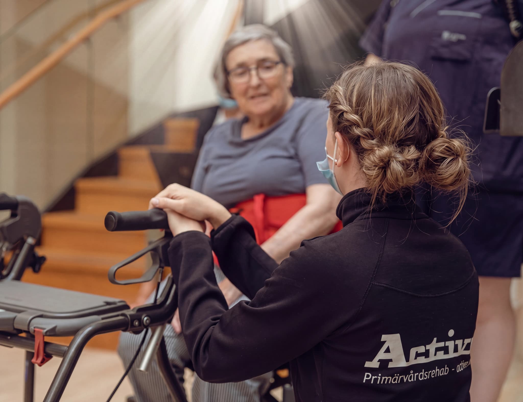 En ung sjukgymnast med flätat hår hjälper en äldre kvinna med glasögon att använda en rollator.