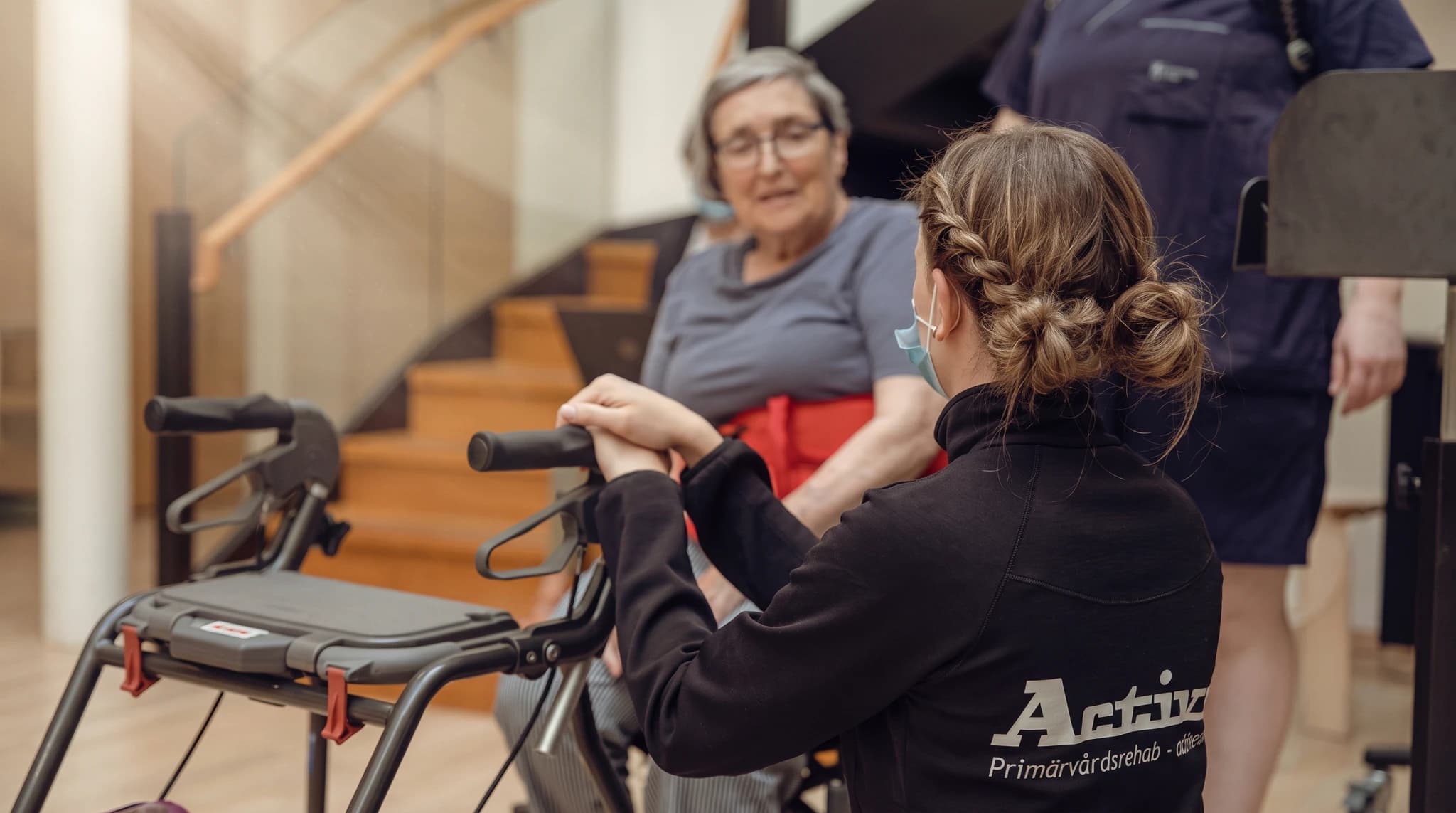 En terapeut med flätat hår hjälper en äldre kvinna med glasögon att använda en rollator.