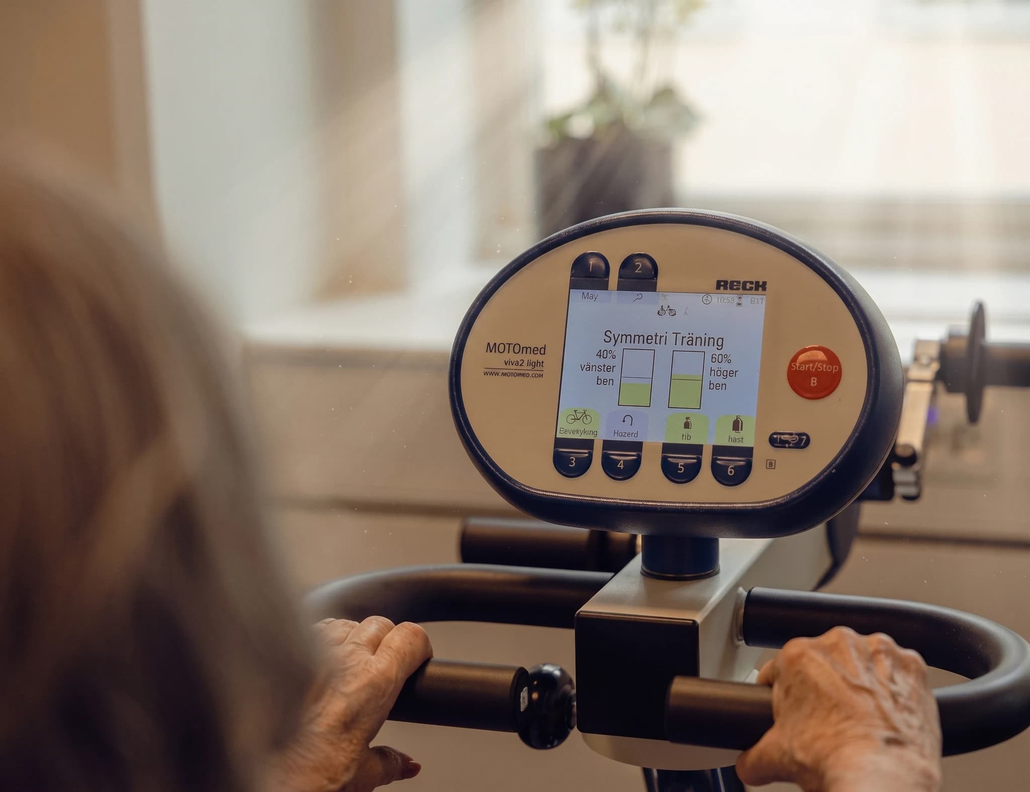 En äldre person använder en MOTOmed motionscykel för rehabilitering med en display som visar symmetrisk träning.