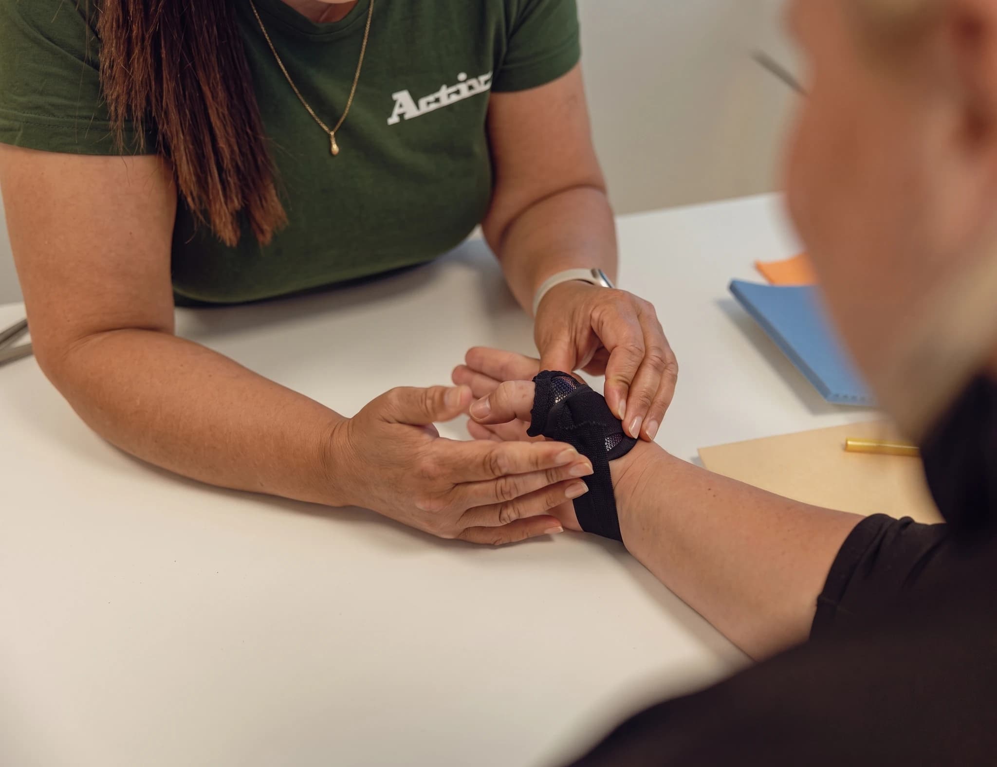 En terapeut applicerar tejpen på handleden på en patient som sitter vid ett bord.