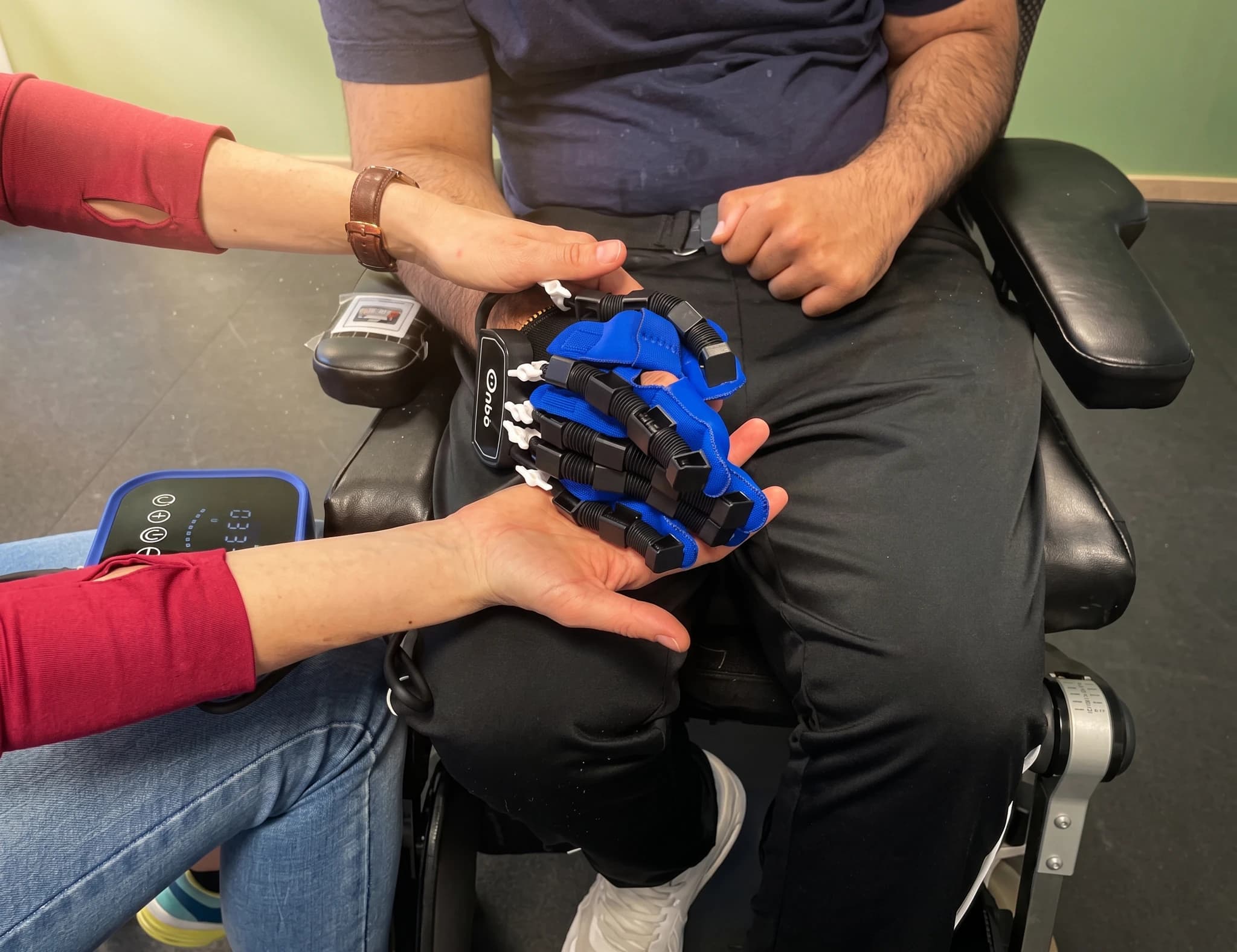 En patient sitter i en rullstol och testar en blå robotisk handhandske, som hjälper till med rehabiliteringsövningar.