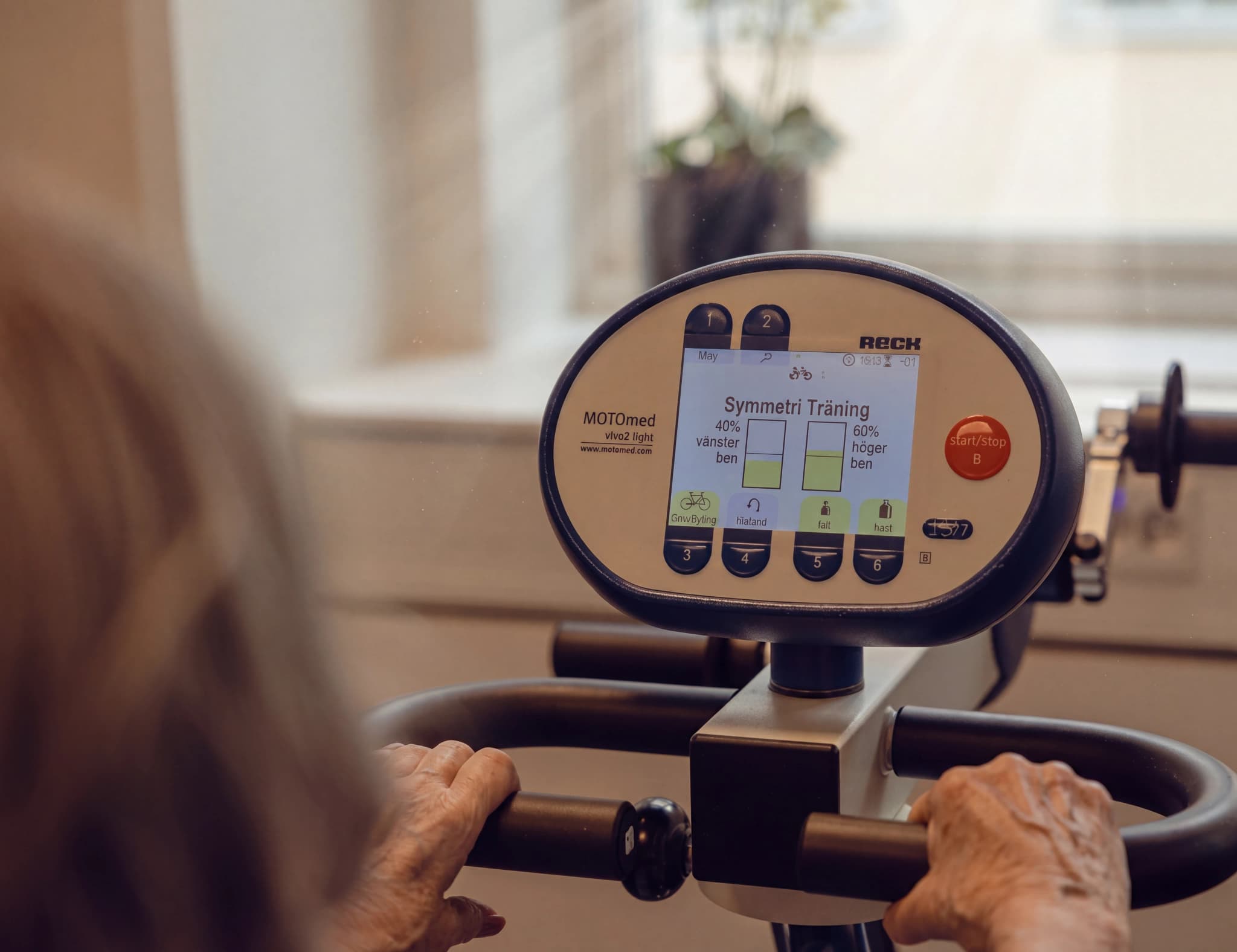 Äldre person som använder en MOTOmed Symmetri Träning rehabiliteringsmaskin med händer på handtagen och tittar på skärmen.