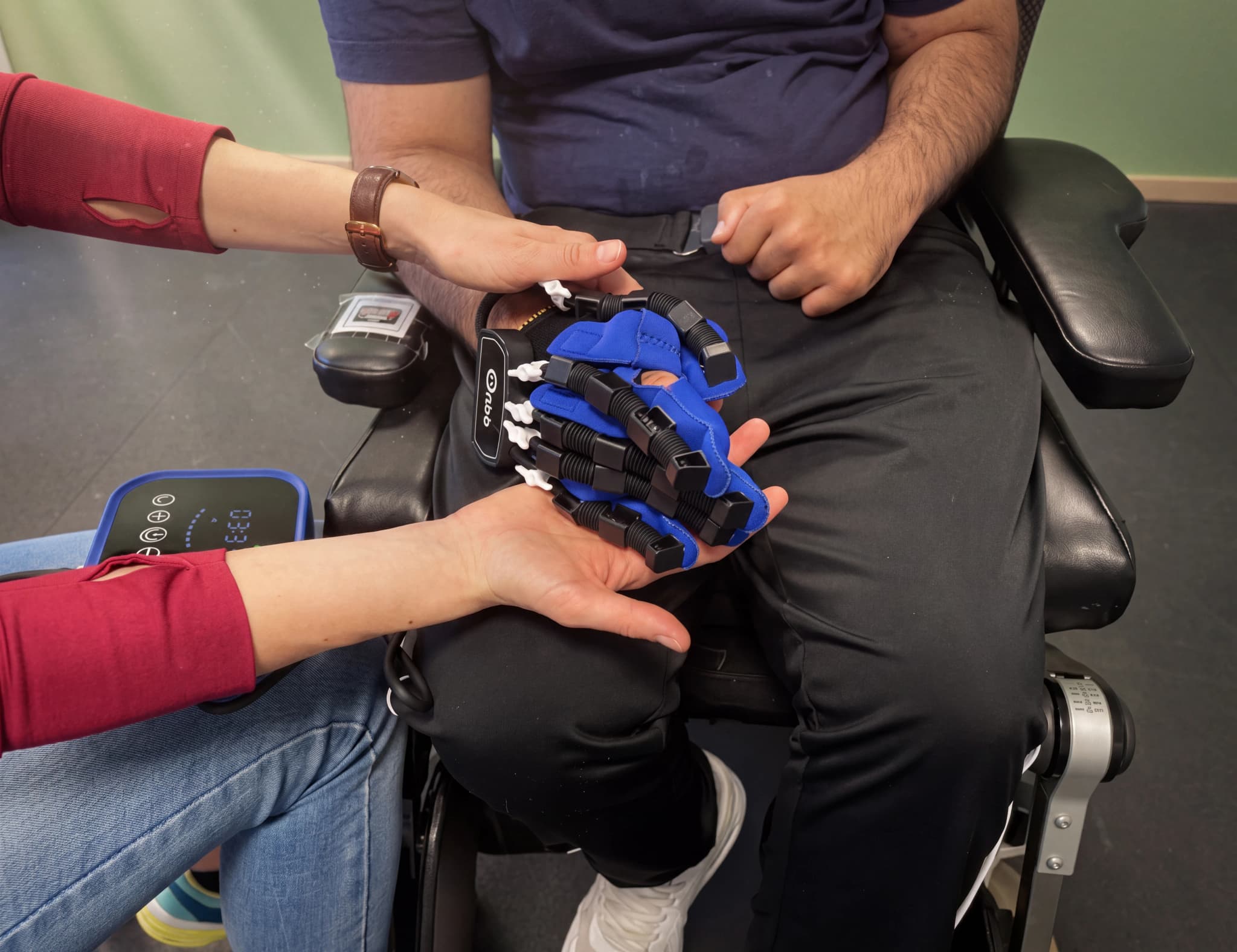 En patient sitter i en stol och använder en blå rehabiliteringshandske med hjälp av en terapeut.