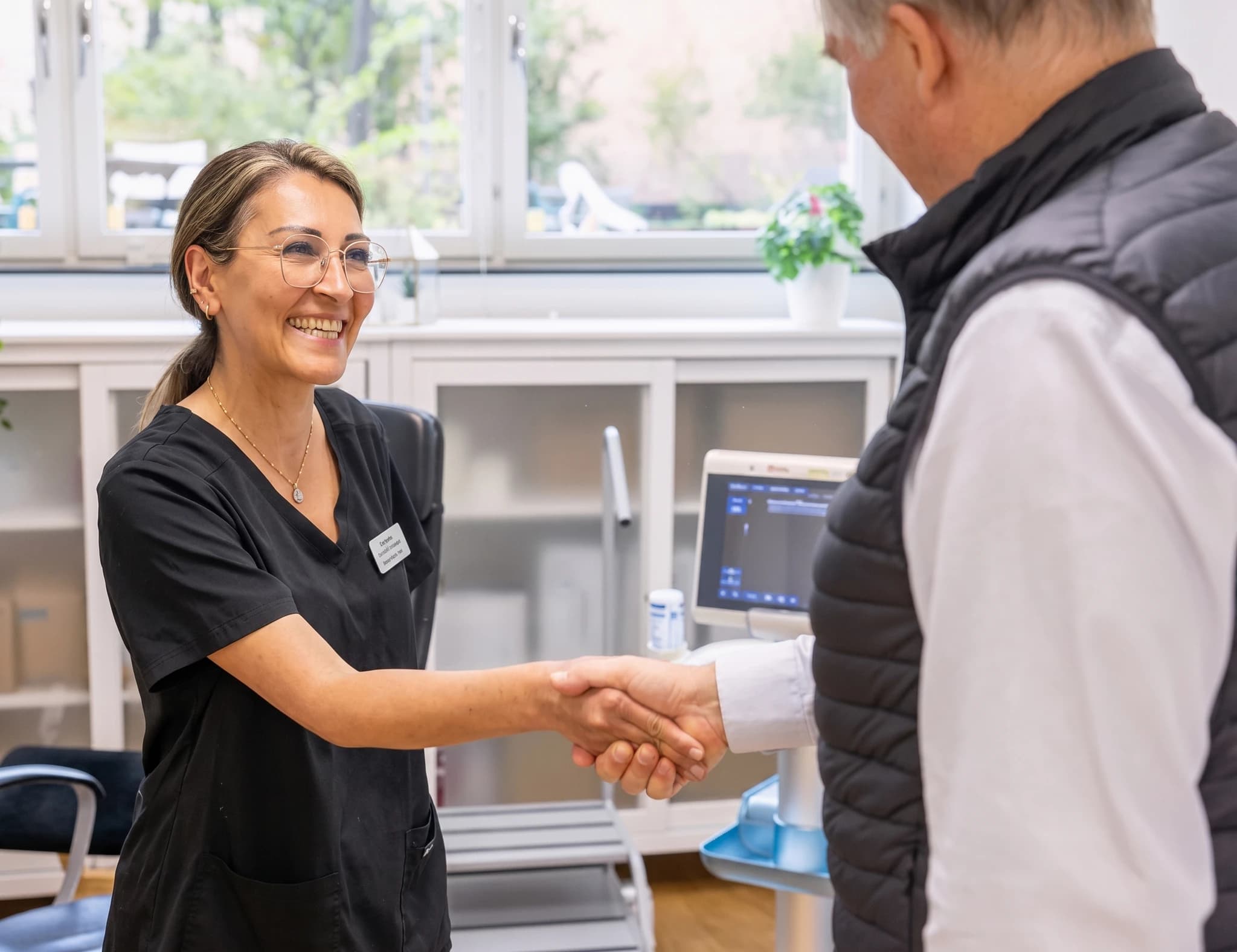 En läkare i mörk scrub-uniform skakar hand med en patient i en klinik, med en ultraljudsapparat i bakgrunden.