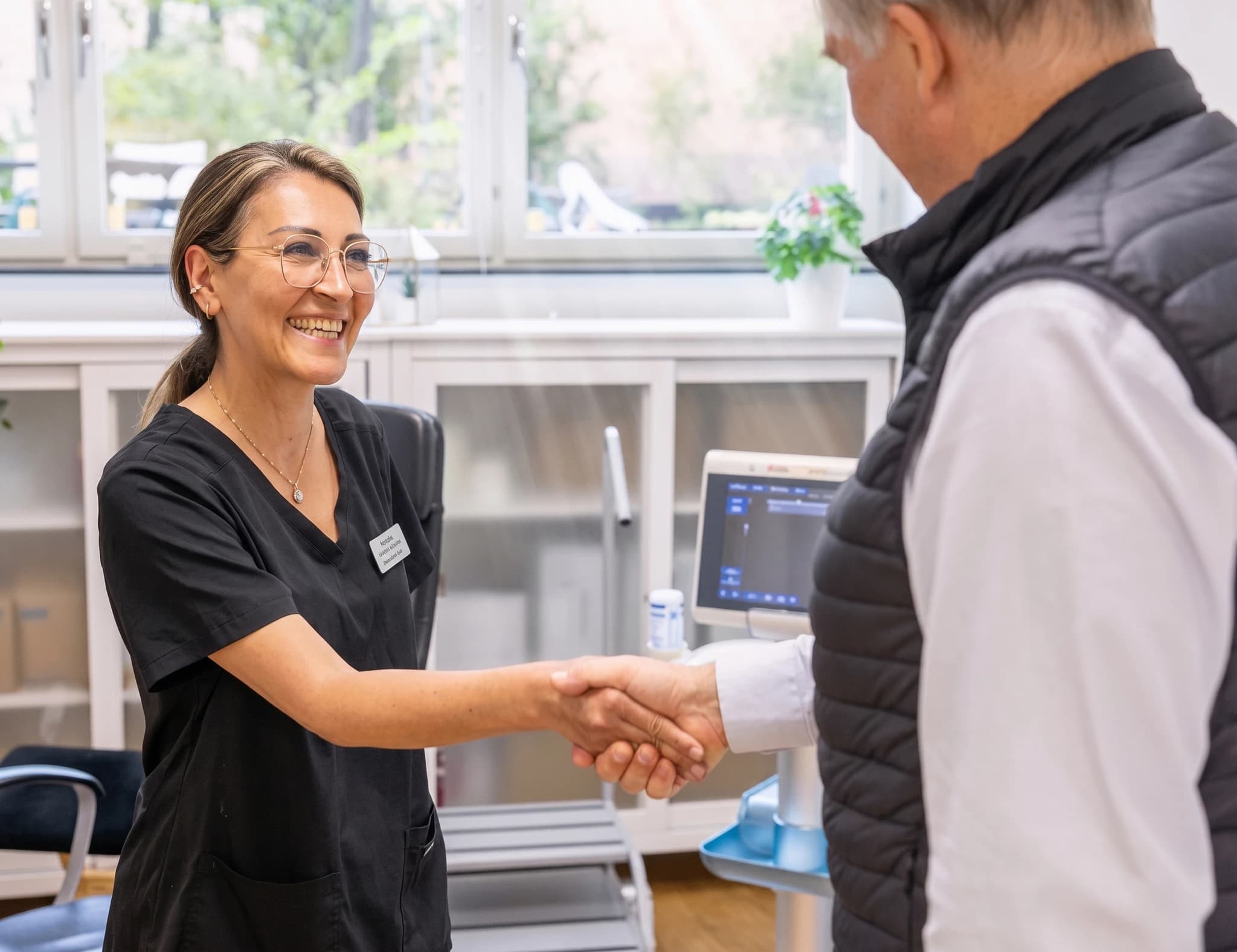 Läkare i svarta scrubs skakar hand med en patient i en klinik, med en ultraljudsmaskin synlig i bakgrunden.