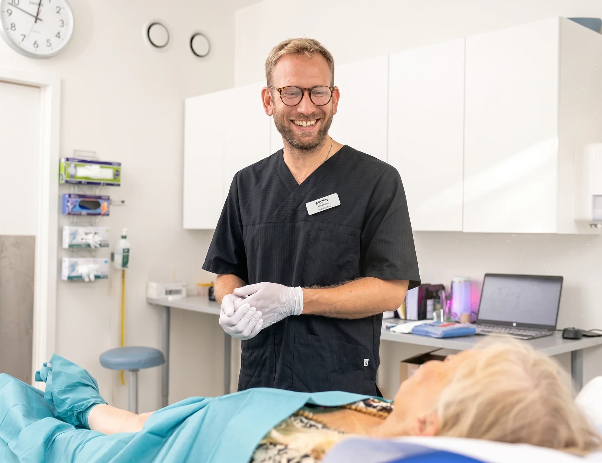 Läkare i mörka kläder med namnbricka pratar med en patient som ligger nedsvept i ett kliniskt rum.