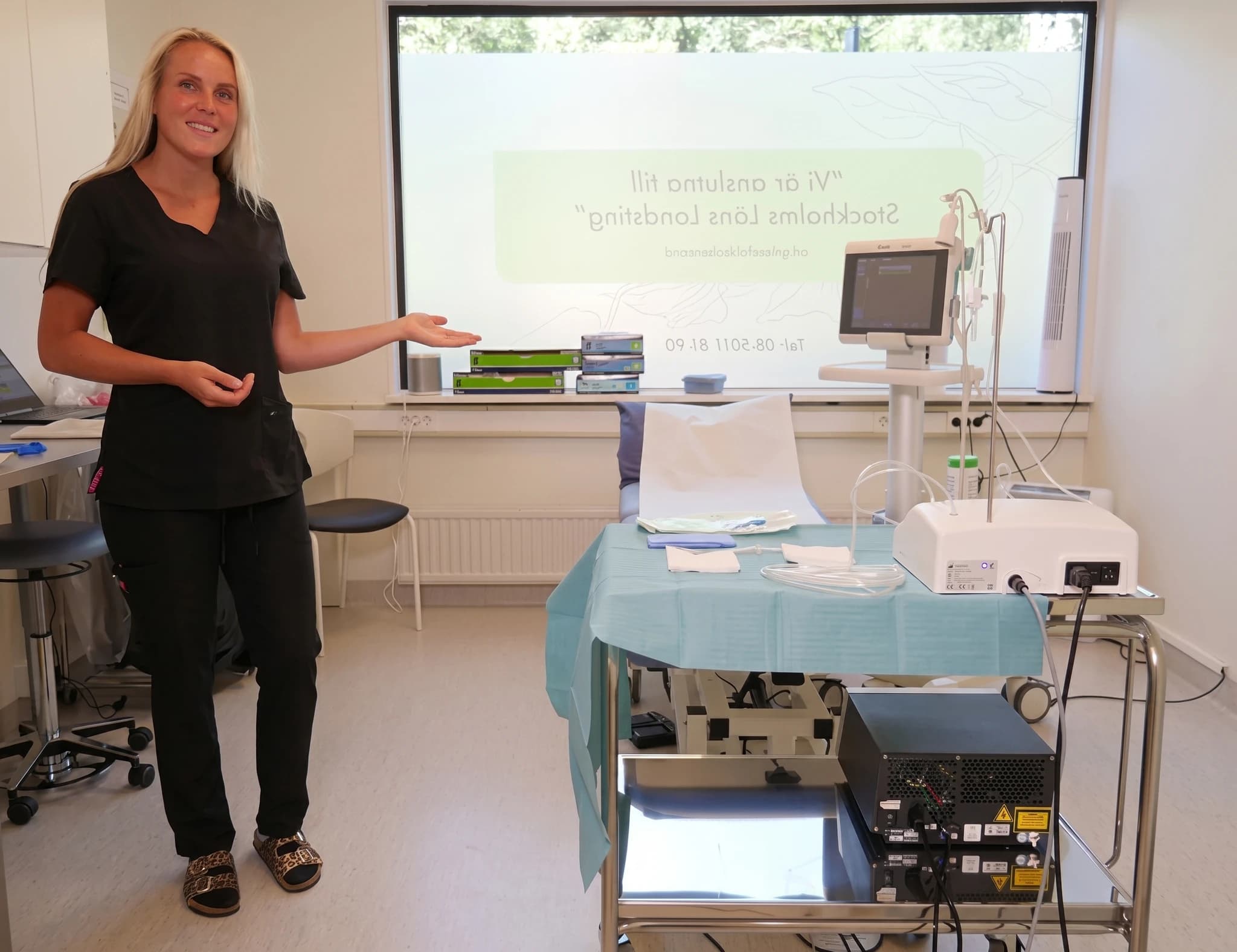En sjukvårdspersonal i mörka kläder pekar mot medicinsk utrustning, inklusive en monitor och en laserapparat, i ett rum.
