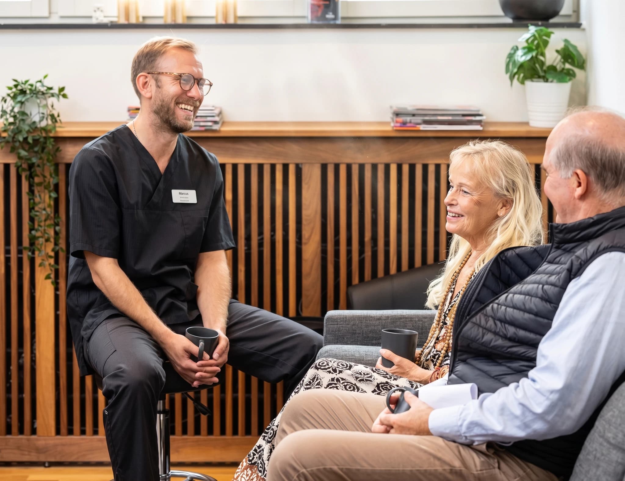 Läkare i svarta arbetskläder ler och pratar med ett äldre par som sitter i en väntrumsmiljö.