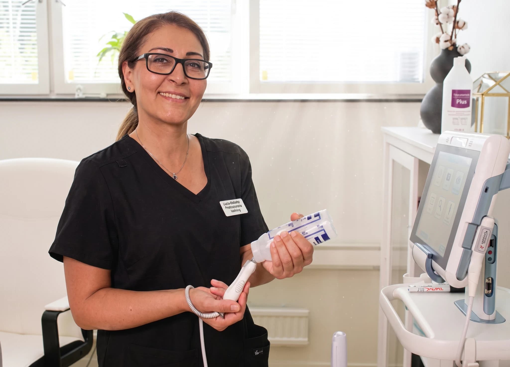 En kvinna i arbetskläder håller upp en medicinsk apparat och ler mot kameran i en klinikmiljö.