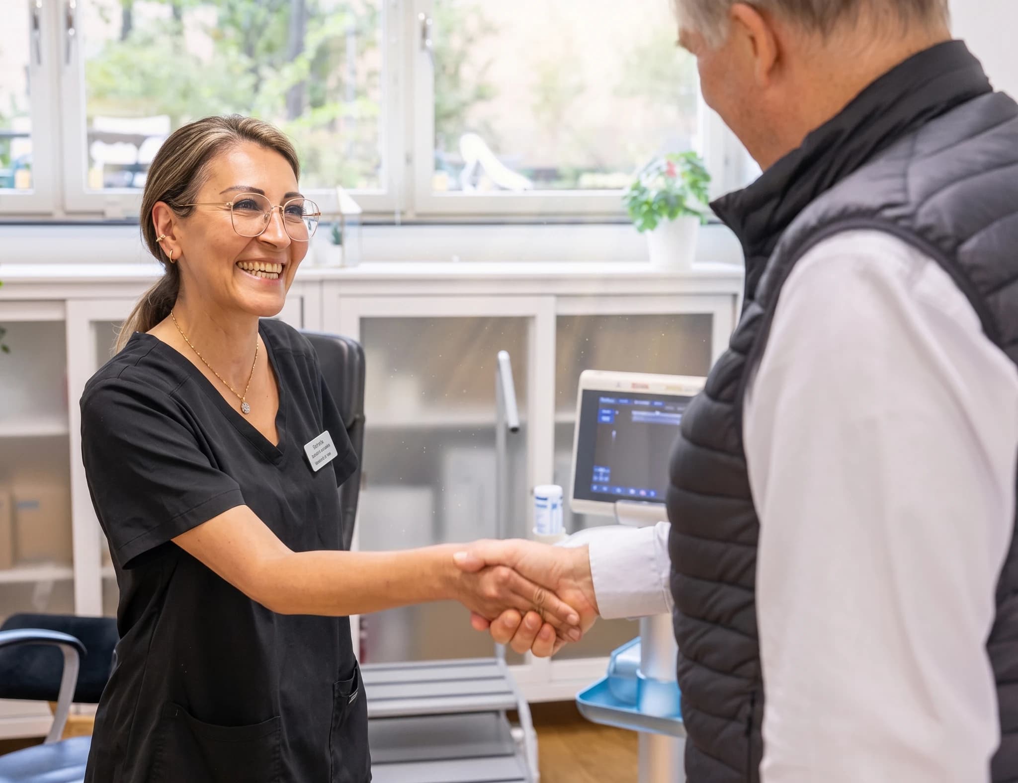 Kvinnlig läkare med glasögon och svart arbetskläder skakar hand med en manlig patient som bär en väst över en vit skjorta, i ett undersökningsrum med medicinsk utrustning.