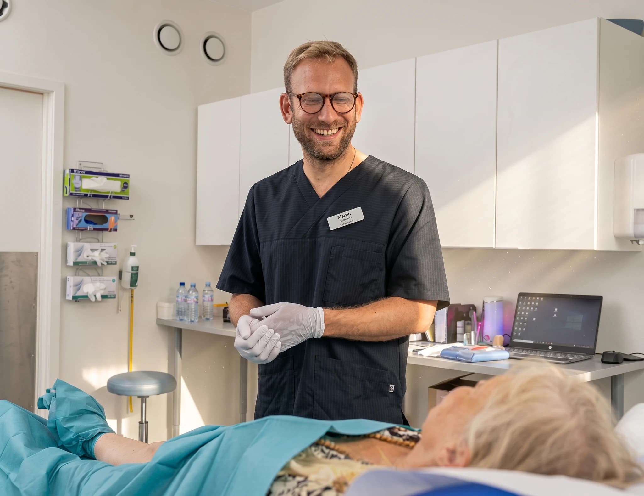 Läkare i mörka kläder och handskar ler medan han tittar på en patient som ligger på en brits, i ett behandlingsrum