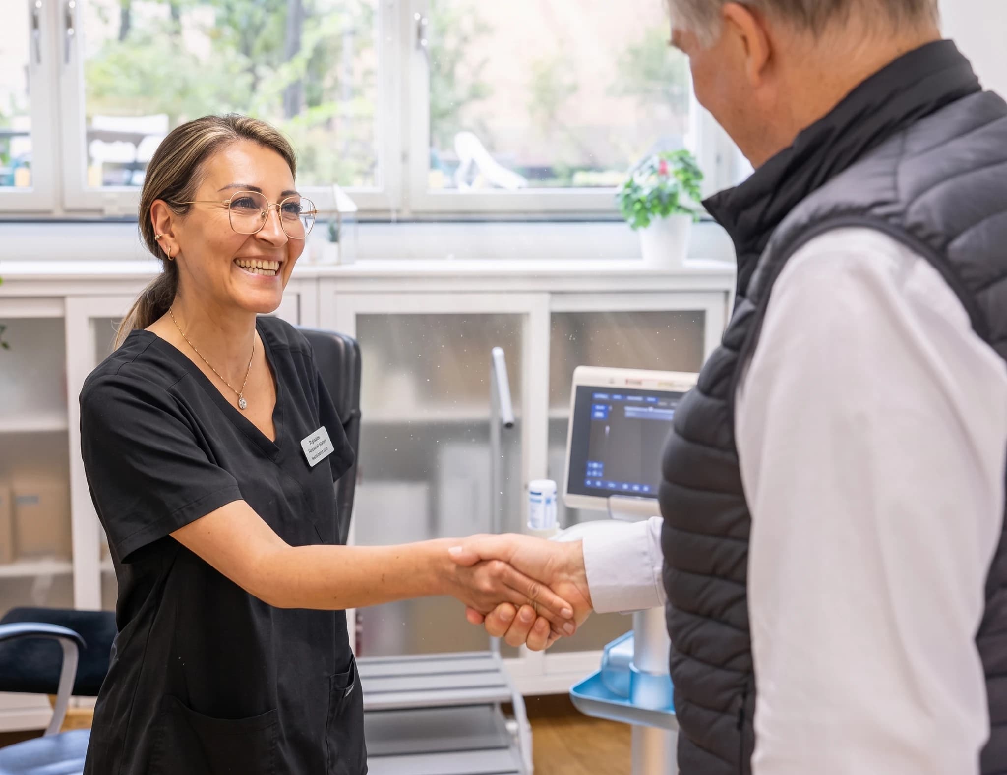 En kvinnlig läkare i svarta arbetskläder skakar hand med en manlig patient i en mottagningslokal.