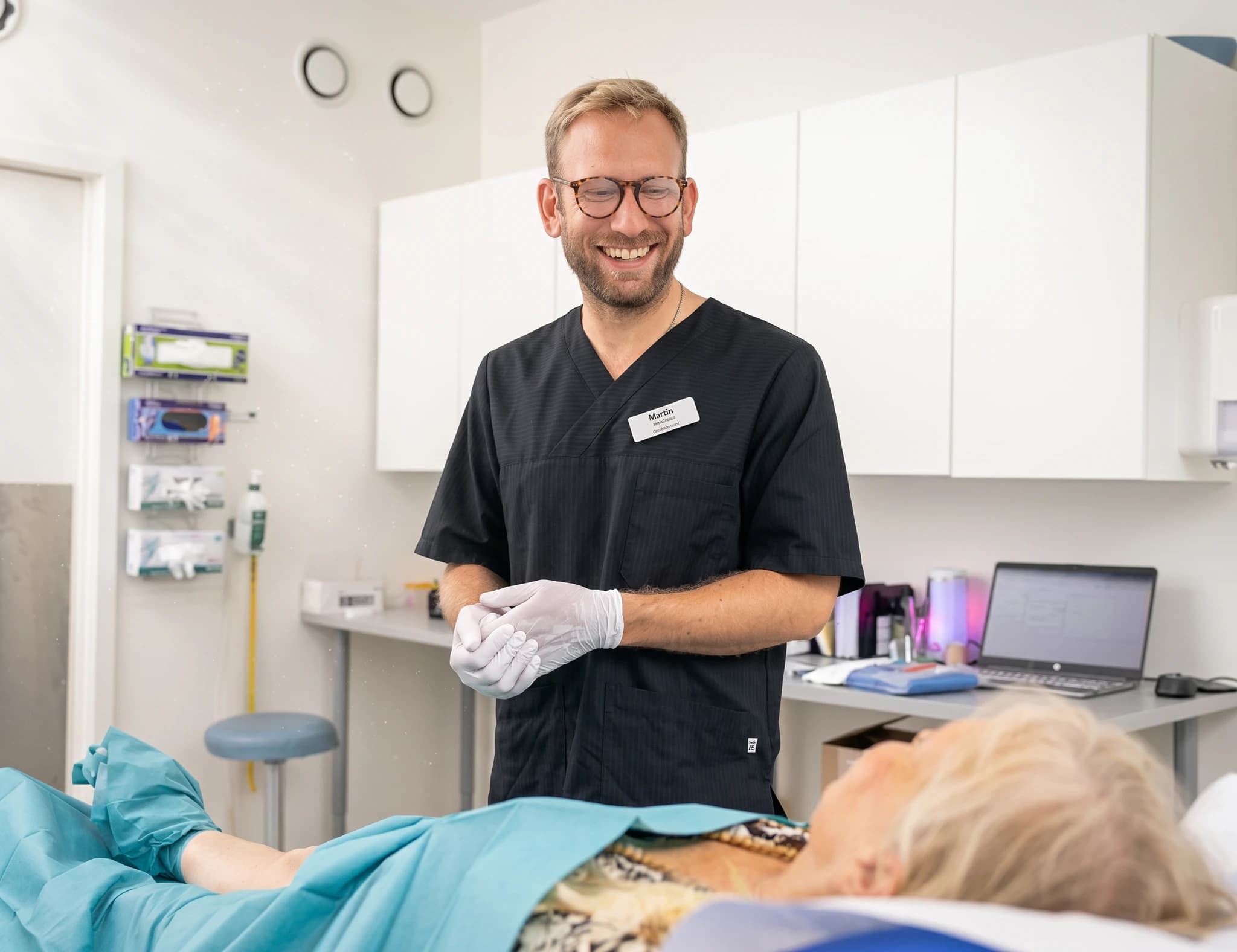 Manlig läkare i läkarrock och handskar pratar med en patient som ligger på en brits i ett undersökningsrum.