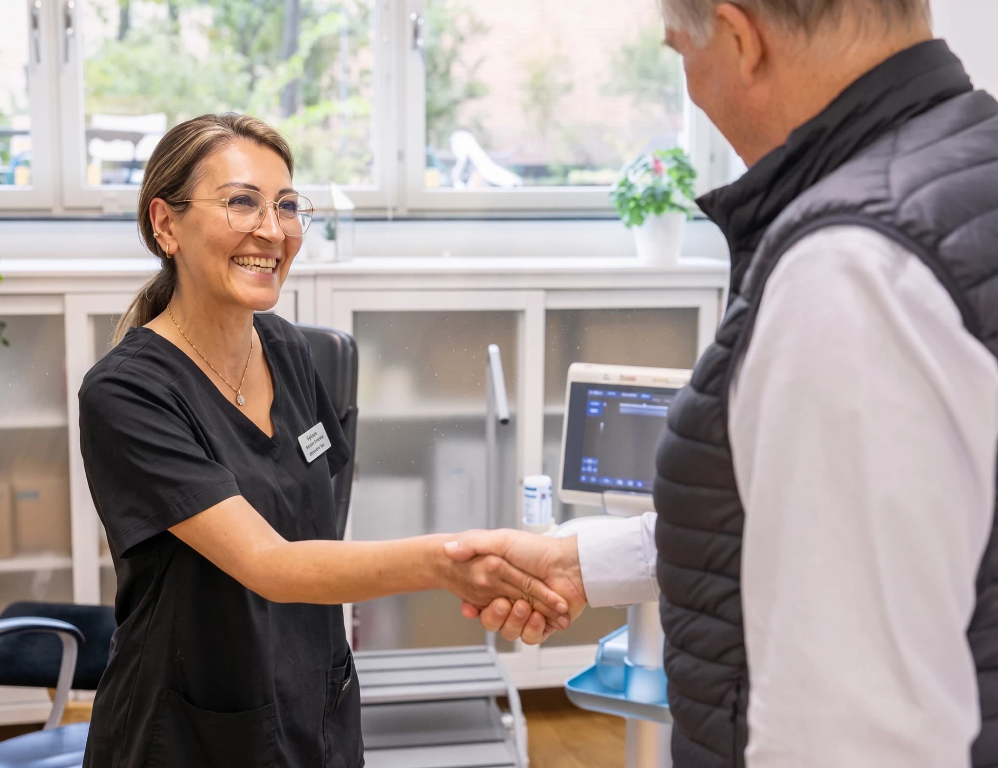 En kvinnlig vårdpersonal med glasögon och svart arbetsklädsel skakar hand med en patient framför medicinsk utrustning på en klinik.