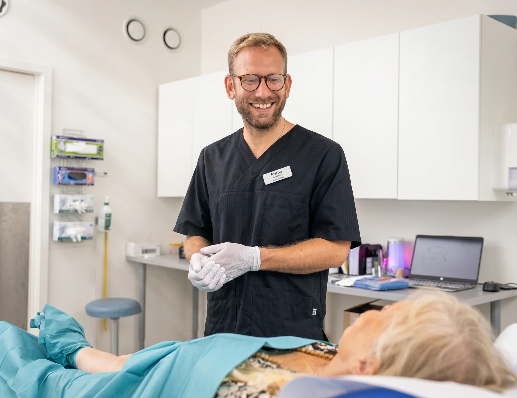 Läkare med glasögon och mörkblå arbetskläder ler medan han håller händer i ett undersökningsrum med en patient täckt av ett grönt draperi.