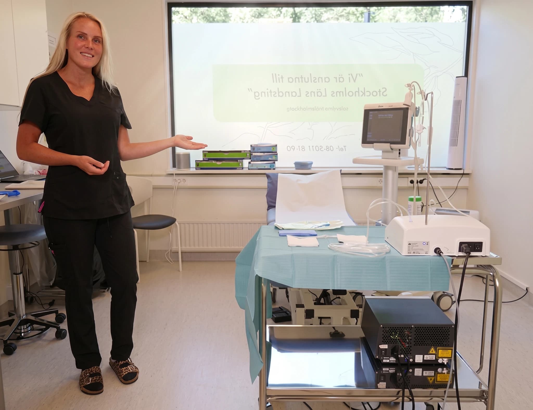 En kvinnlig vårdgivare i arbetskläder gestikulerar och presenterar medicinsk utrustning i ett undersökningsrum.