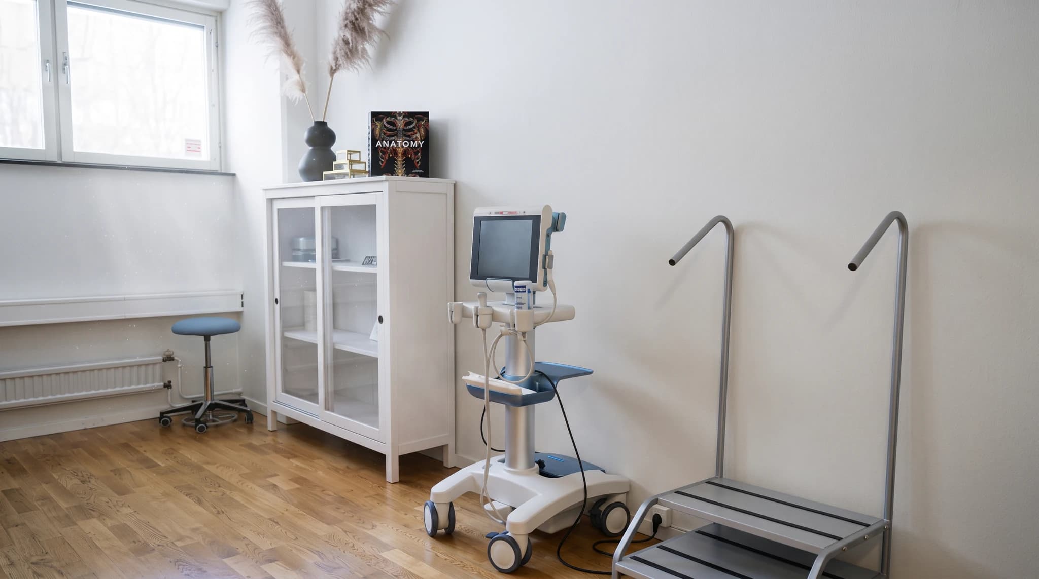 Utrustning för medicinsk undersökning, inklusive en ultraljudsmaskin och ett skåp med glasdörrar, i ett kliniskt rum.