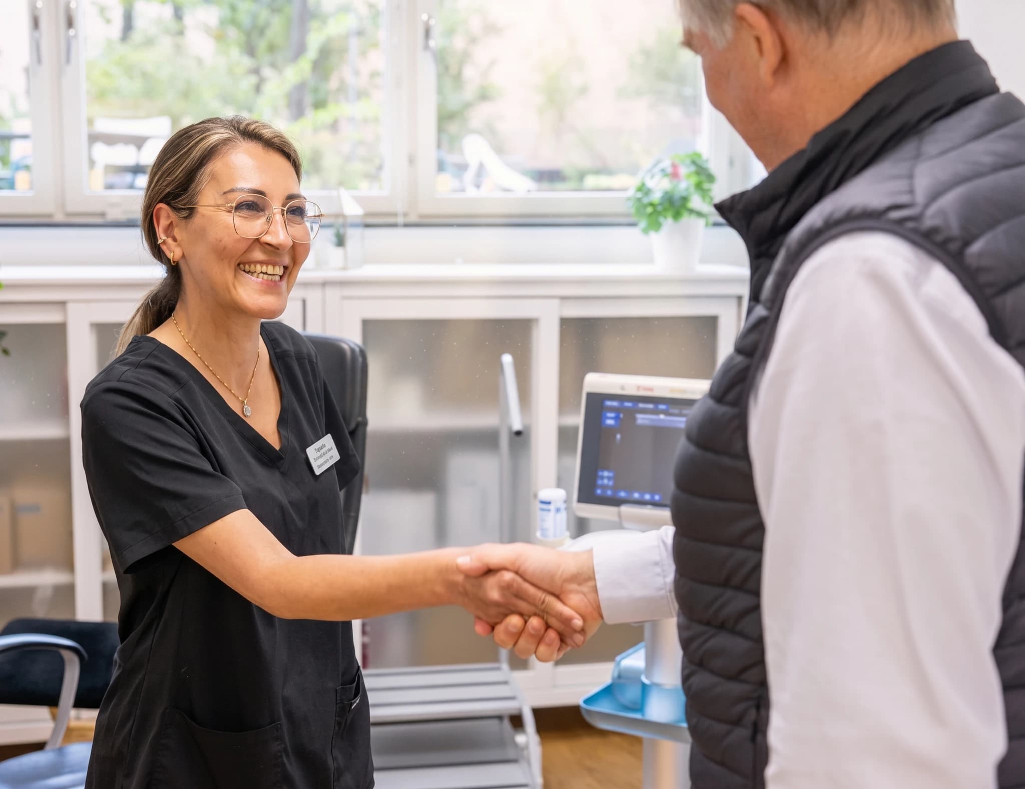 Läkare i mörk scrubs skakar hand med en man i väst framför en ultraljudsmaskin.