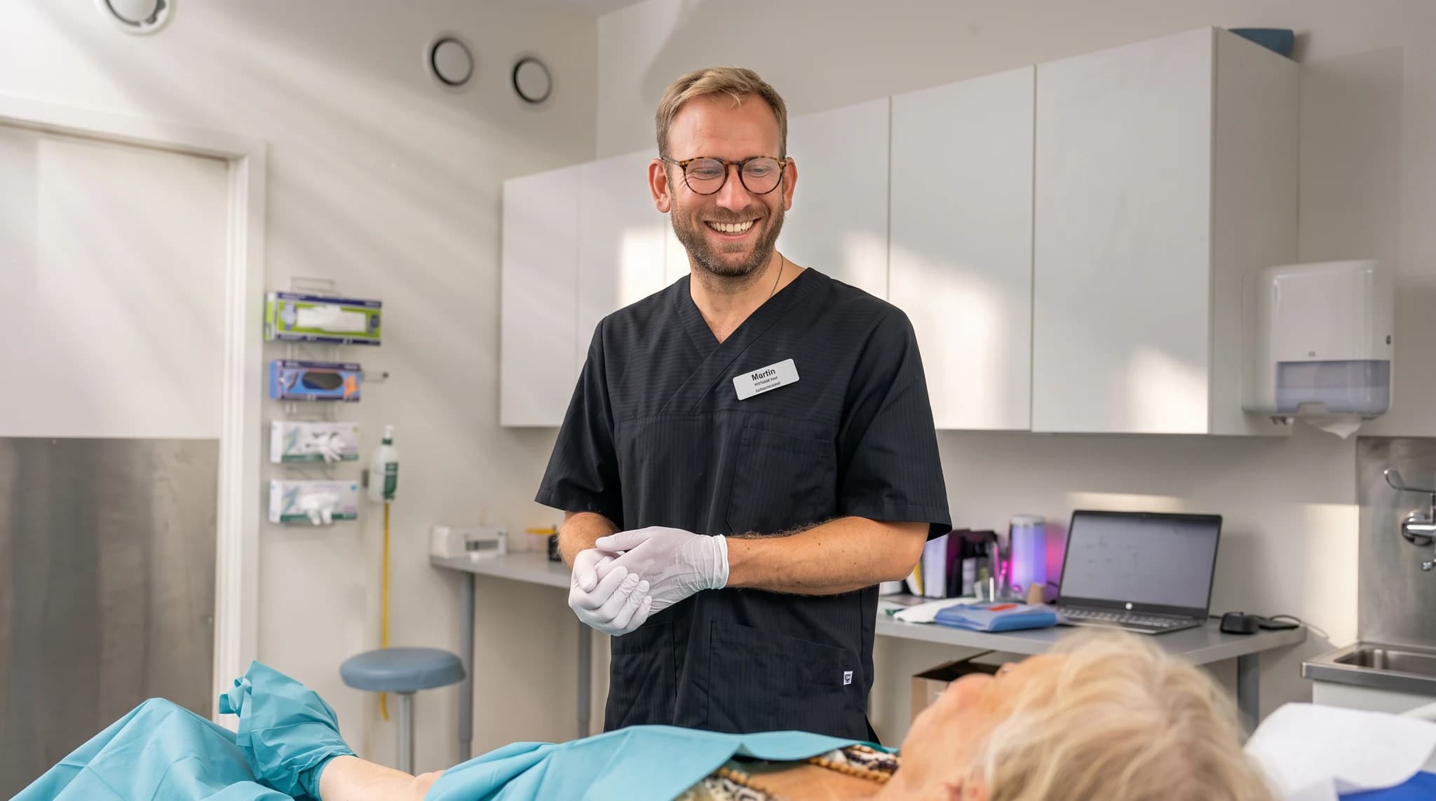 Läkare i svarta kläder, med glasögon och handskar, ler mot en patient som ligger på en brits täckt med gröna dukar i ett kliniskt rum.