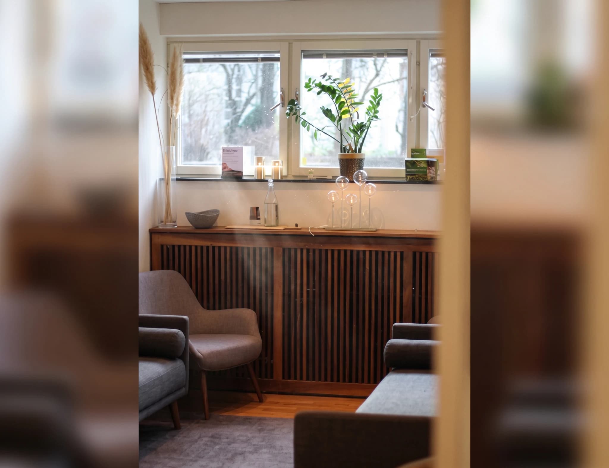 Väntrum med gråa fåtöljer, en låg träbänk med ribbad front och en fönsterbräda med växter och dekorativa föremål.