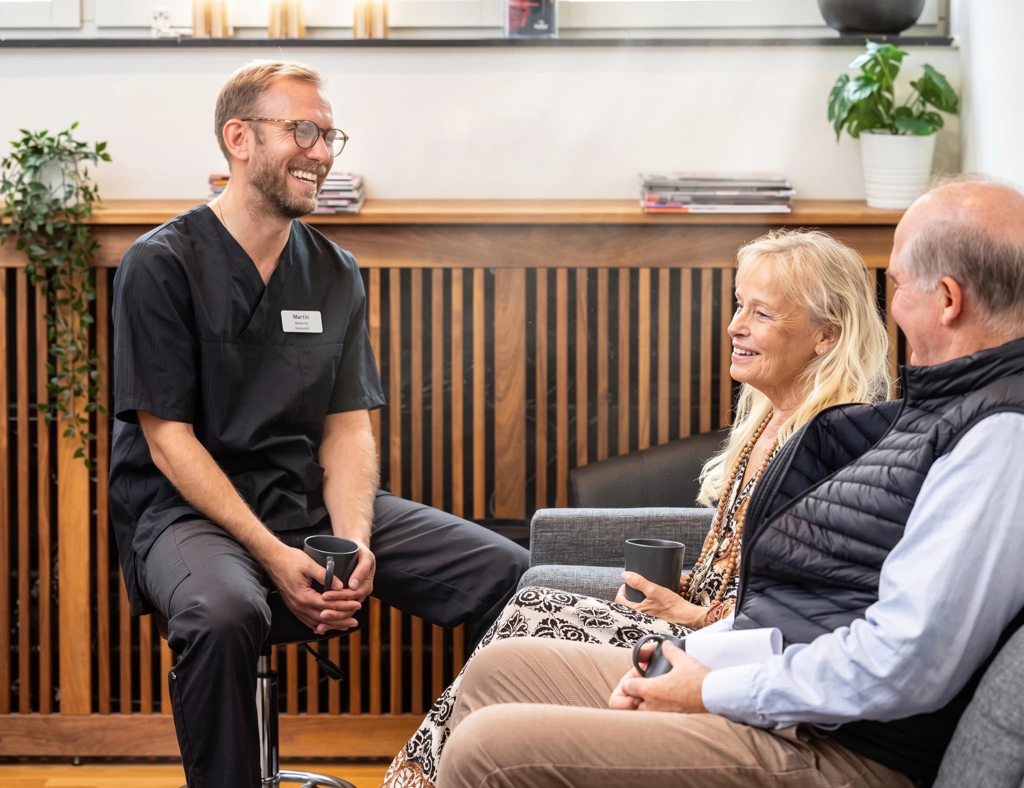 Tandläkare i svarta arbetskläder sitter och pratar med ett äldre par som sitter i en väntrumsmiljö med kaffe.