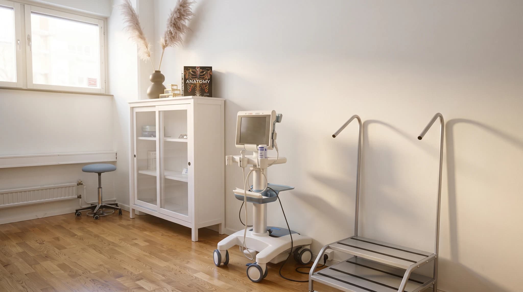 Klinikrum med ett ultraljudsmaskin, ett vitt skåp med glasdörrar, en undersökningsstol och två stegpallar.