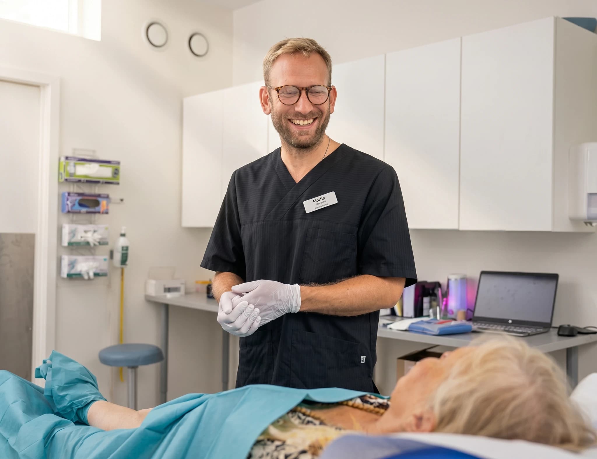 Läkare i svarta kläder med handskar som ler och pratar med en patient i ett behandlingsrum.