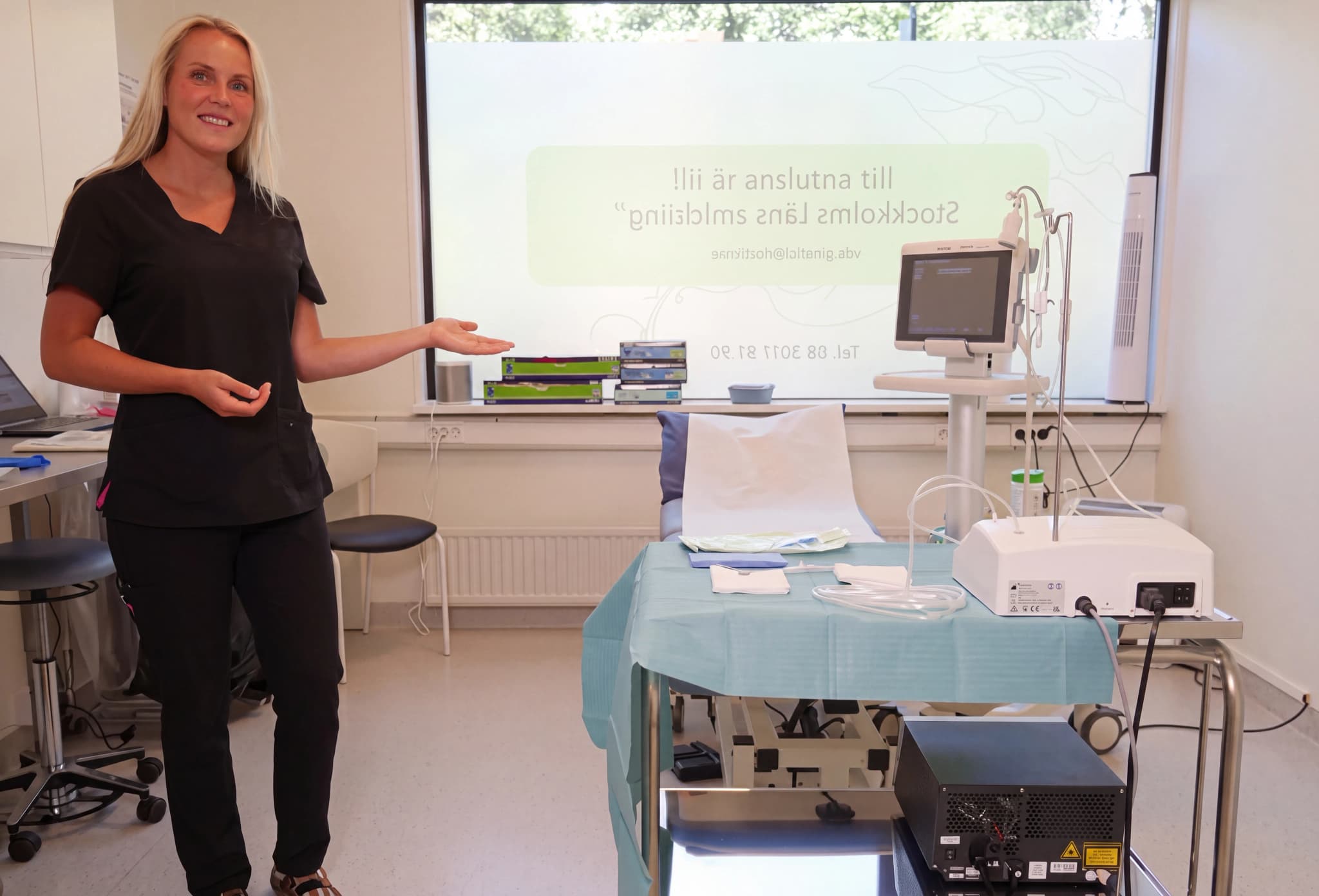 En sjukvårdspersonal i mörkblå arbetskläder presenterar medicinsk utrustning framför en stor skärm i ett undersökningsrum.