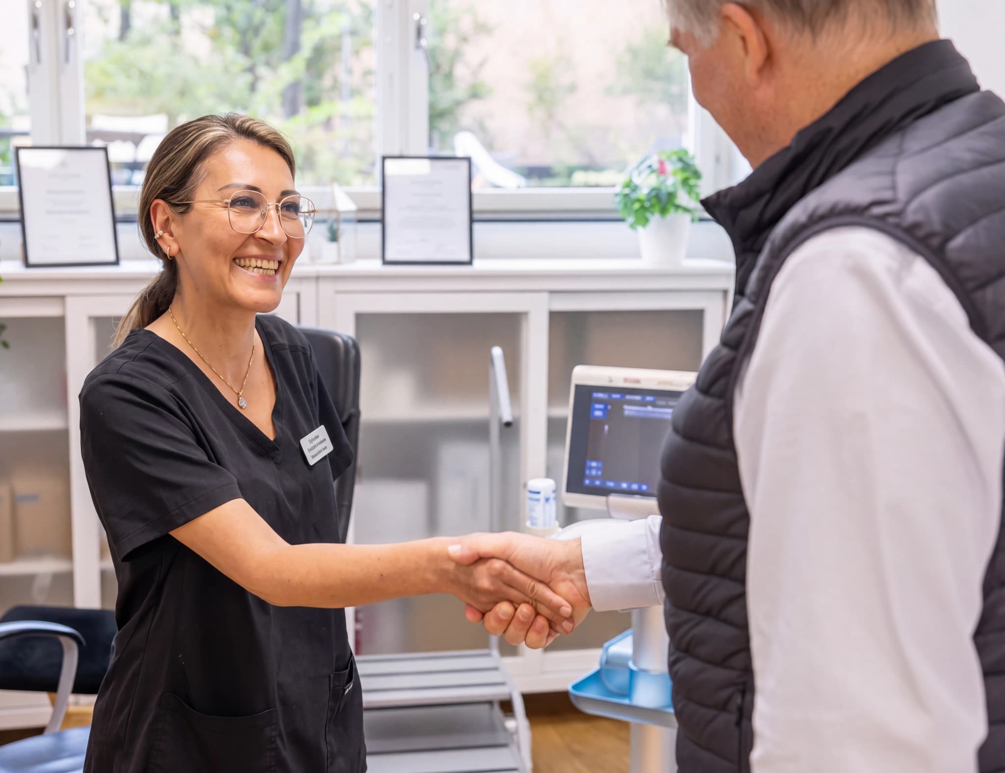 En kvinnlig vårdpersonal i svarta scrubs ler och skakar hand med en patient framför medicinsk utrustning.