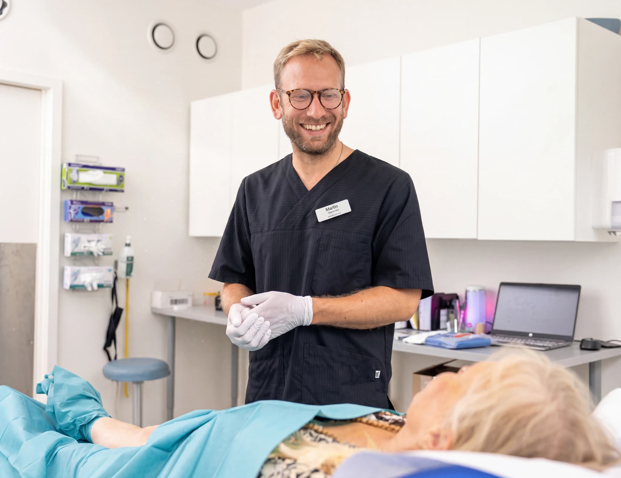 Läkare Martin med glasögon och svart läkarrock ler medan han håller händerna i vita engångshandskar, bredvid en patient som ligger på en undersökningsbrits i ett ljust undersökningsrum.