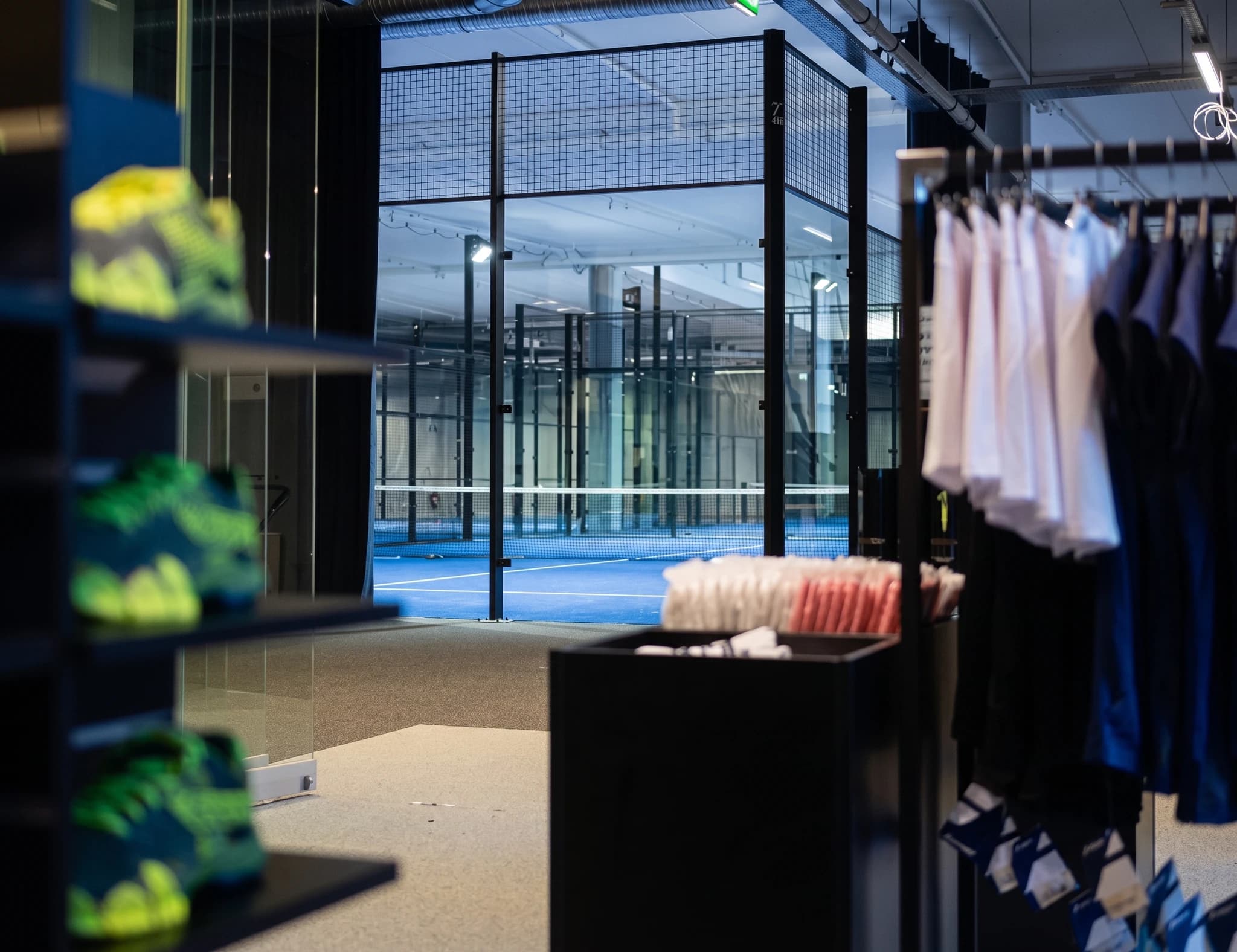 En butik med sportutrustning, inklusive skor och kläder, med en inomhuspadelarena med blå bana i bakgrunden.