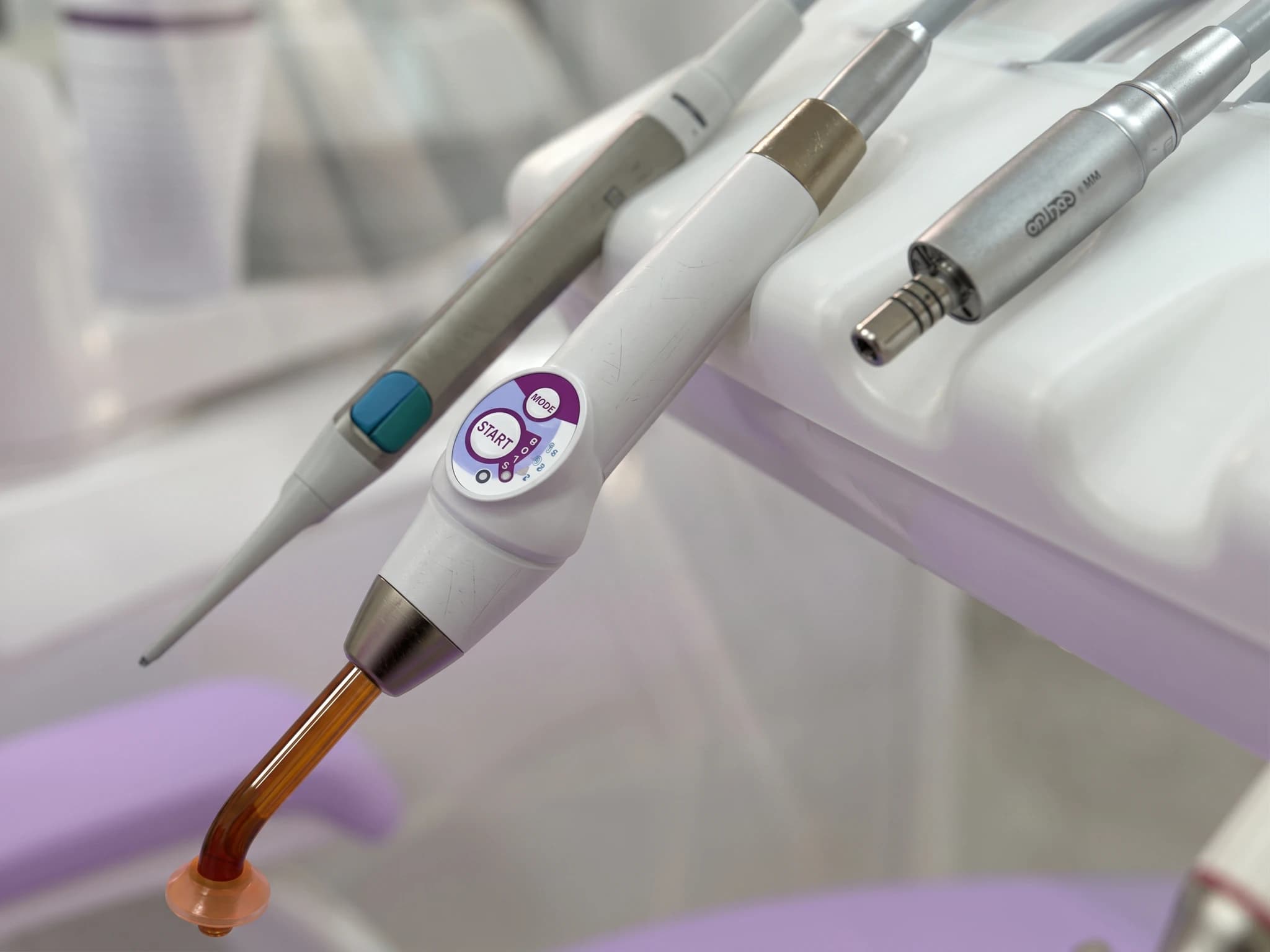 En närbild på en vit och silverfärgad tandvårdsljus härdningsenhet med en droppe på spetsen, bredvid andra tandläkarinstrument i en klinik.