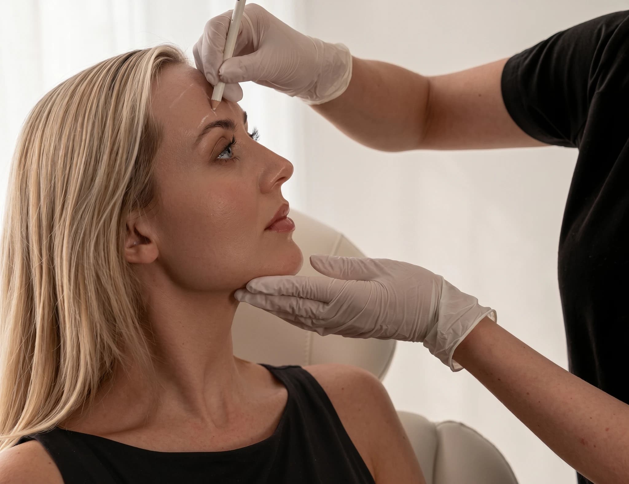 Estetiker markerar patientens panna med en penna innan behandling, patienten sitter i en stol.