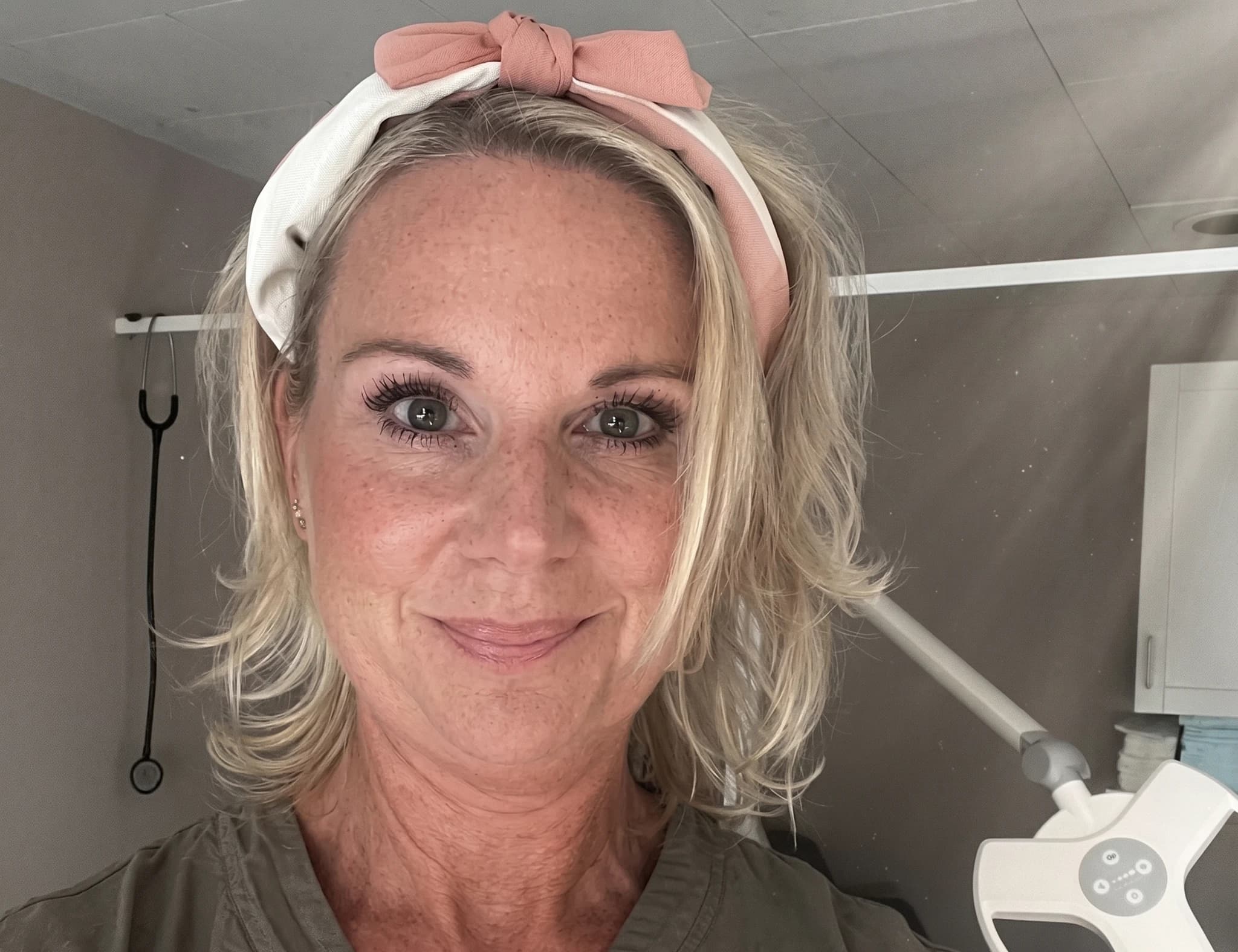 Kvinna med pannband ler mot kameran i ett klinikutrymme, med stetoskop och medicinsk utrustning i bakgrunden.