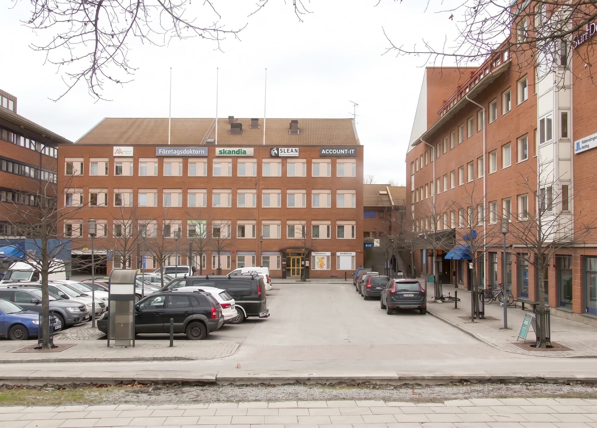 Exteriör av en tegelstensbyggnad med flera våningar, som rymmer olika företag. En parkeringsplats med flera bilar syns framför byggnaden under en molnig dag.