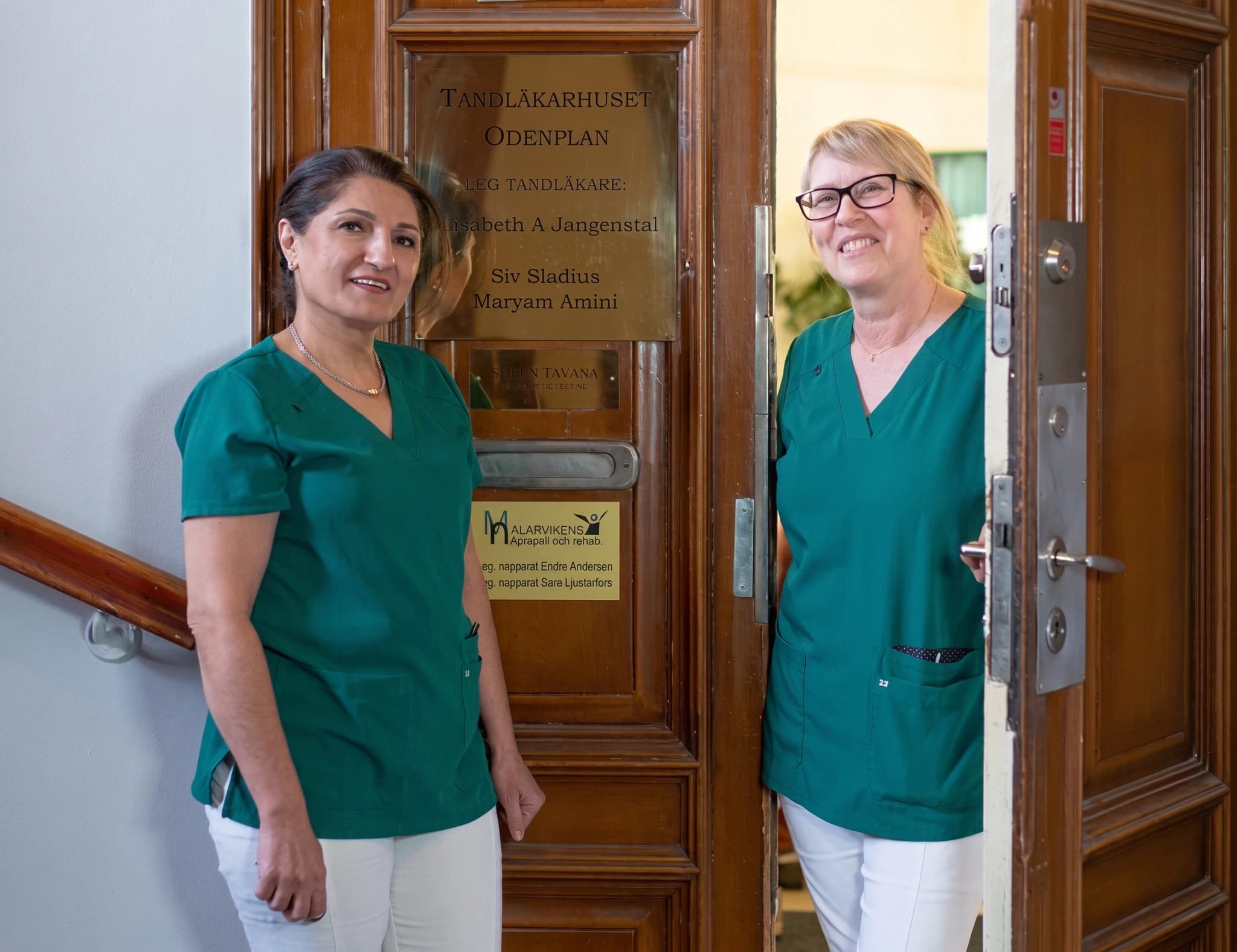 Två tandläkare, en klädd i grön tunika och en i ljusblå tunika, ler vänligt vid ingången till en tandläkarklinik.