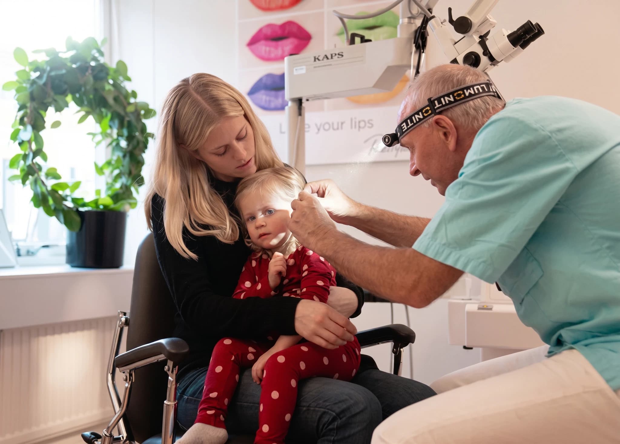 Läkare undersöker ett barns öra med ett otoskop i ett undersökningsrum, med en mamma som håller barnet.