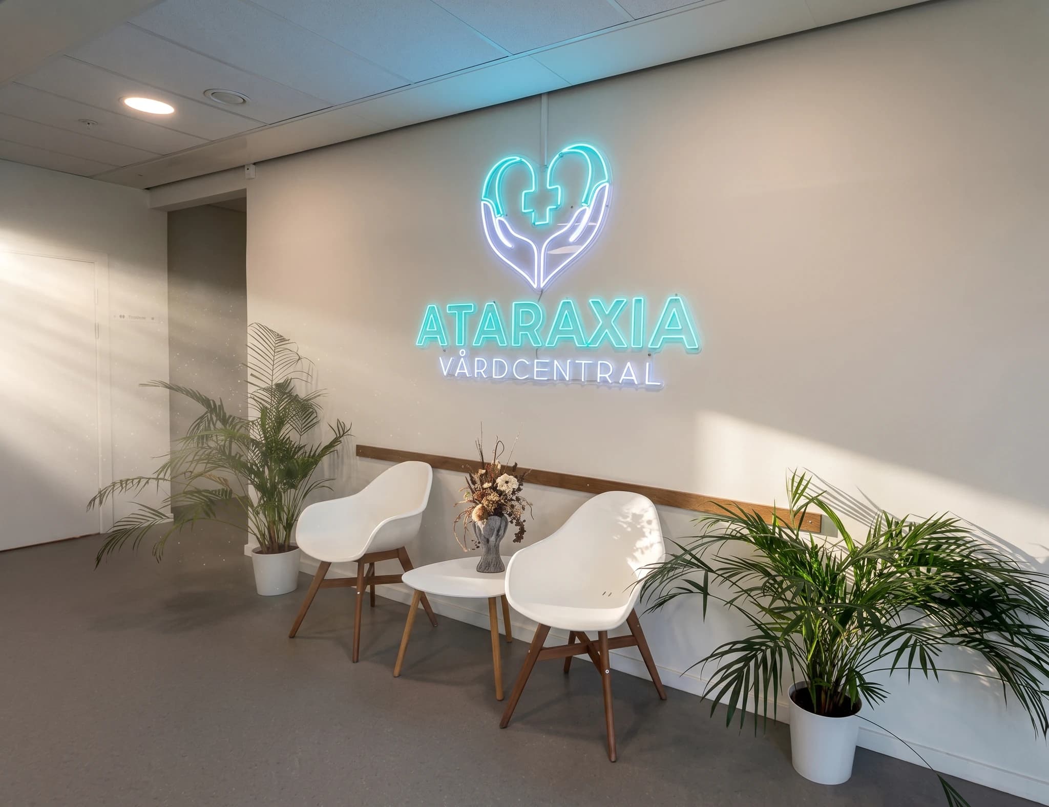 Ataraxia Vårdcentral neonskylt med hjärt- och handmotiv ovanför en väntplats med två vita stolar och ett litet bord med blommor.