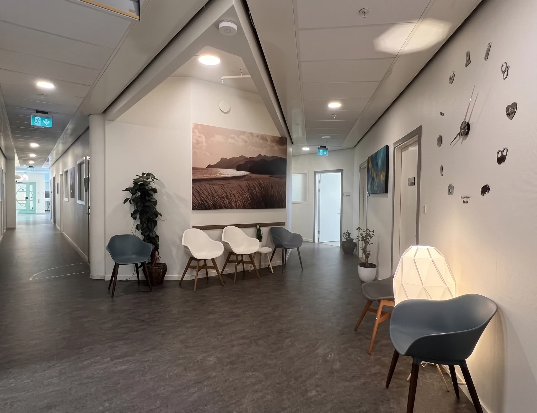 En väntrumskorridor med sittplatser, en stor väggklocka och konstverk.
