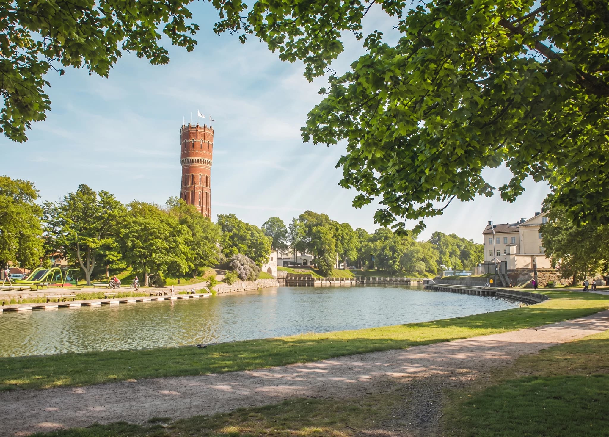 En vacker vy av ett vattentorn i tegel med en kronliknande topp, beläget intill en lugn flod. Parkområdet framför vattentornet har grönskande träd och en stig. En klassisk byggnad syns till höger om floden.