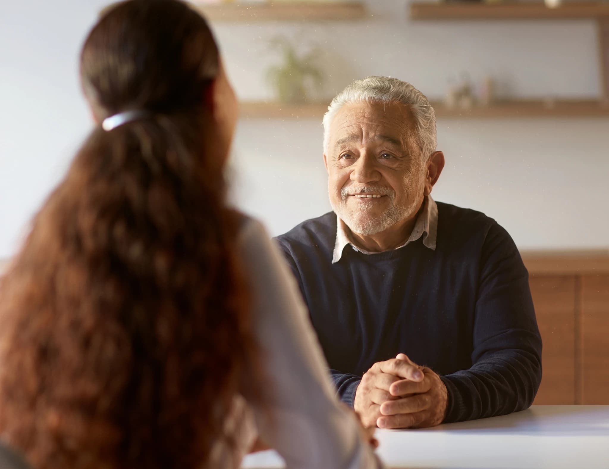 Äldre man med grått hår och ett leende tittar på en person som sitter framför honom.
