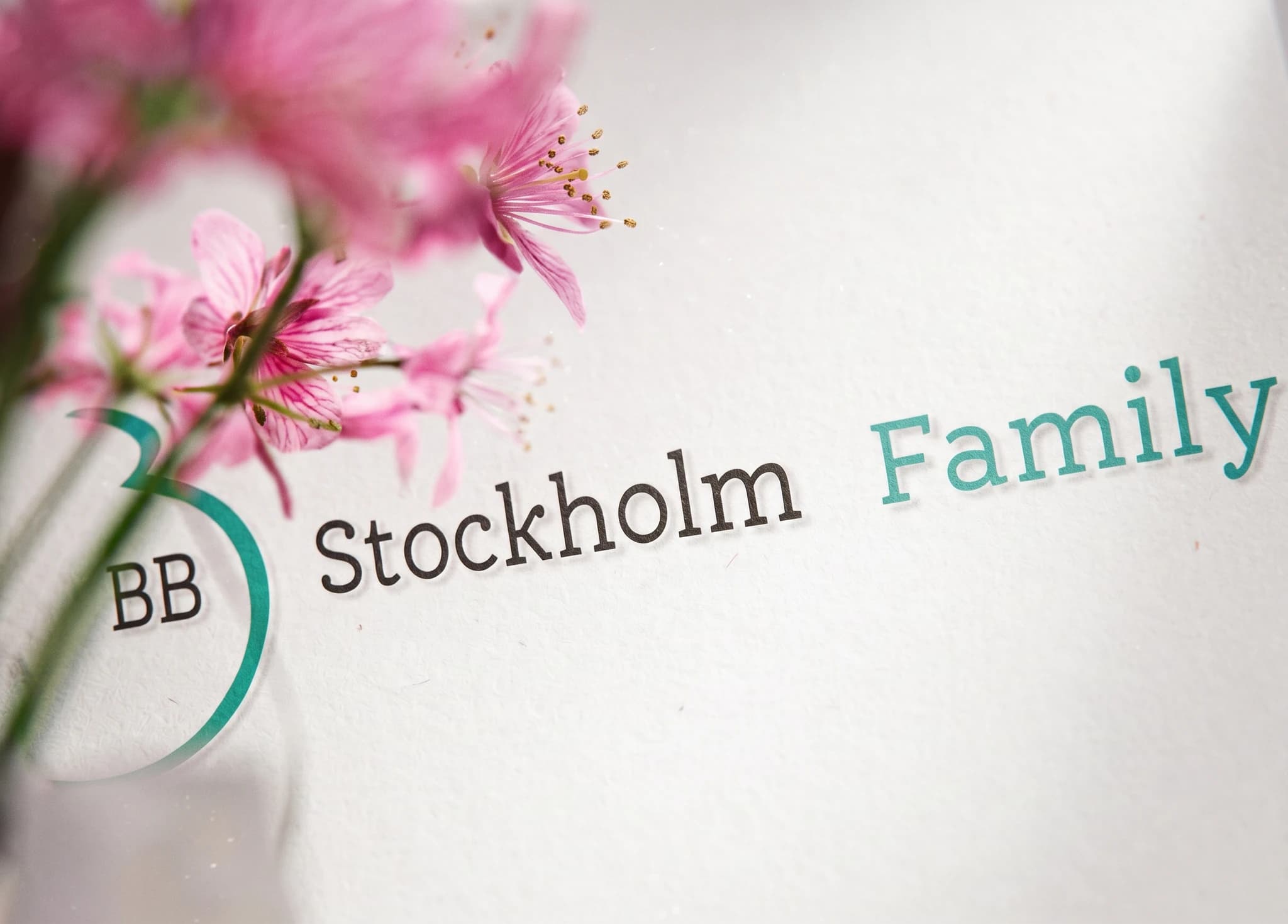 Grafiskt element med texten 'BB' i en cirkel, 'Stockholm' och 'Family' över en suddig bakgrund av rosa blommor.