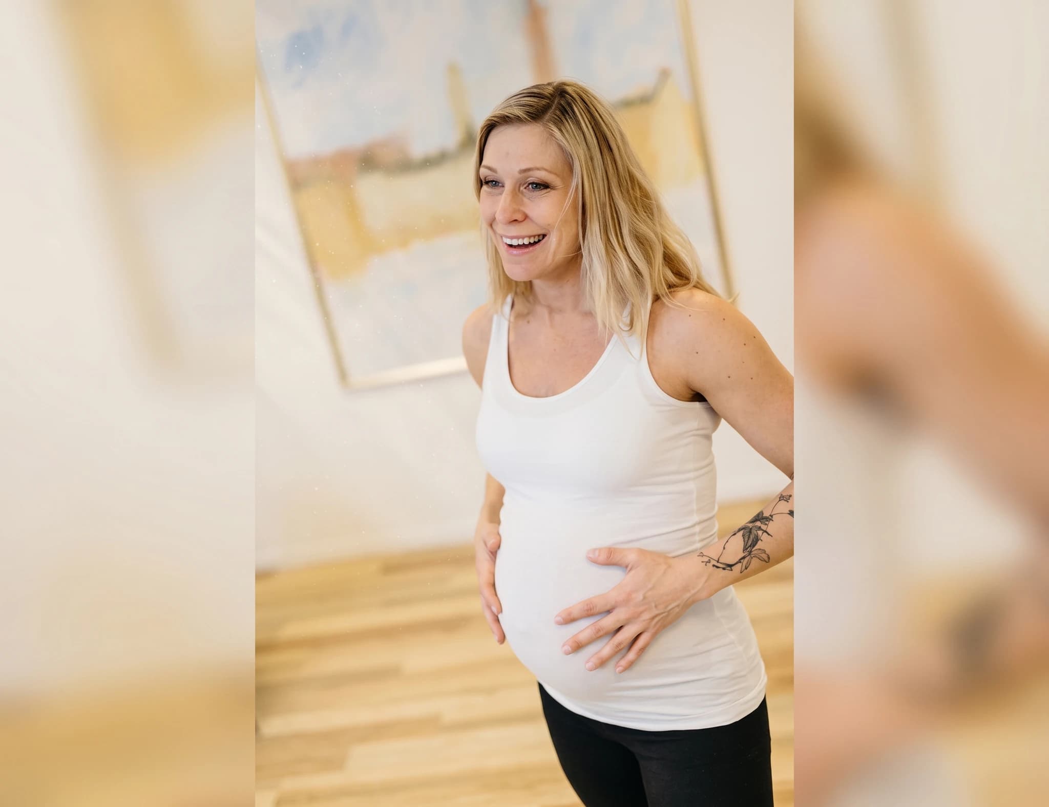 Gravid kvinna med blont hår som ler och håller sig om magen i ett rum med trägolv och en tavla på väggen.