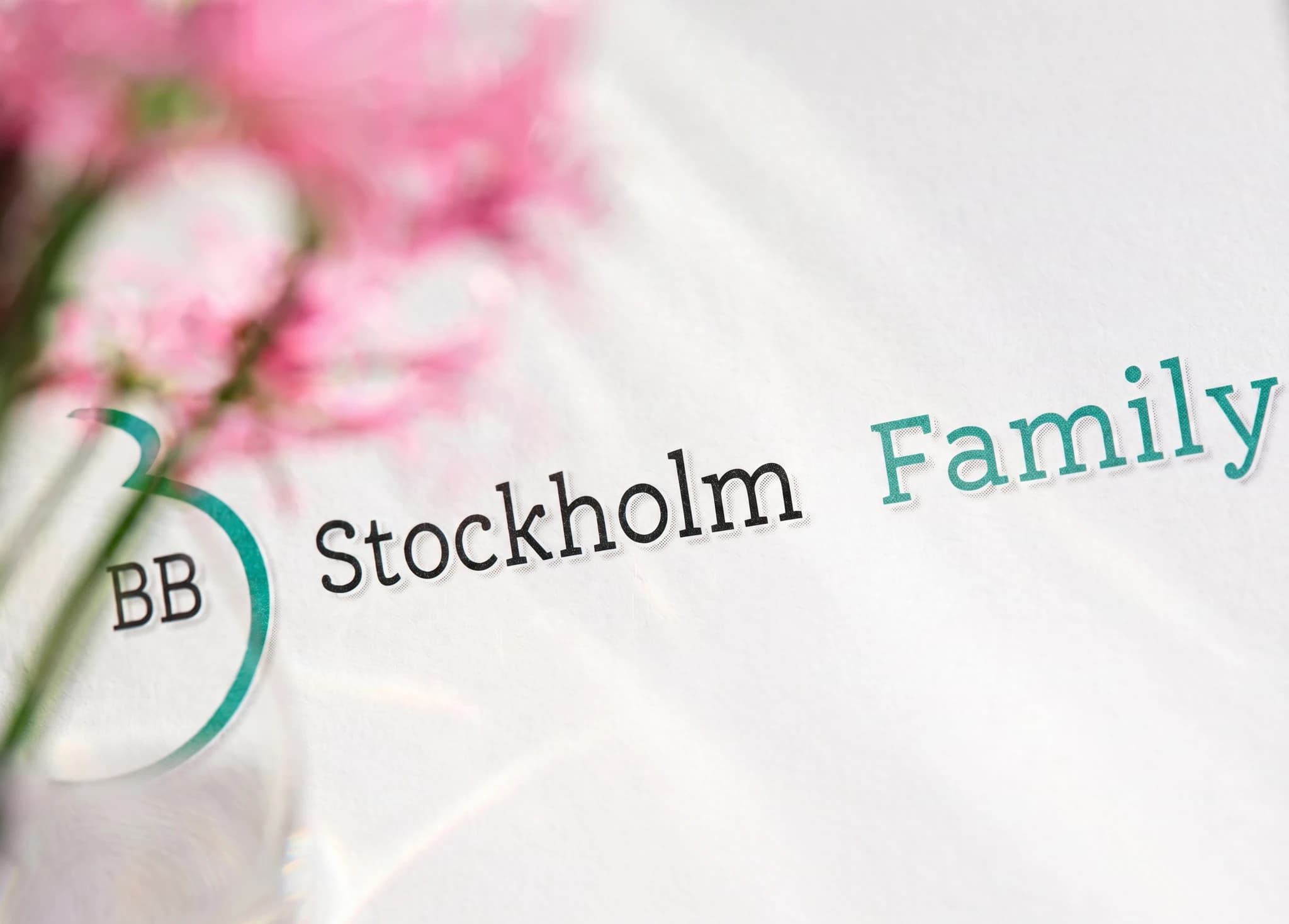 Ett foto av texten "Stockholm Family" på ett vitt papper med en oskarp bakgrund av rosa blommor. Till vänster finns en cirkel med "BB" inuti.