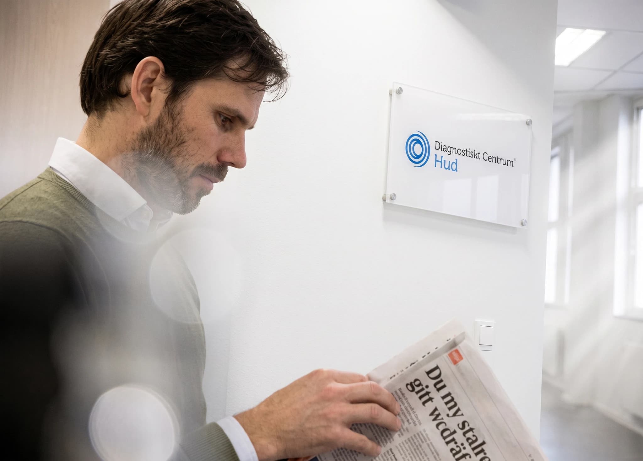 Man i kostym som läser en tidning framför en väggskylt för Diagnostiskt Centrum Hud.